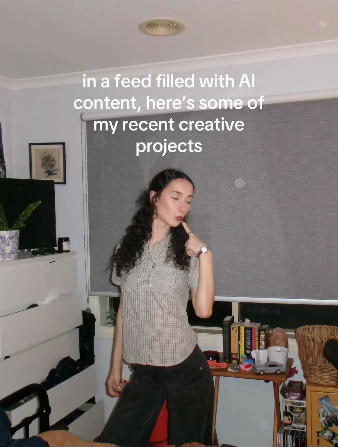 A woman with curly dark hair and light skin standing in a room, touching her lips with her finger. She is wearing a gray checkered shirt and black pants, with a watch on her wrist. The room contains a television, books, and various decorative items.