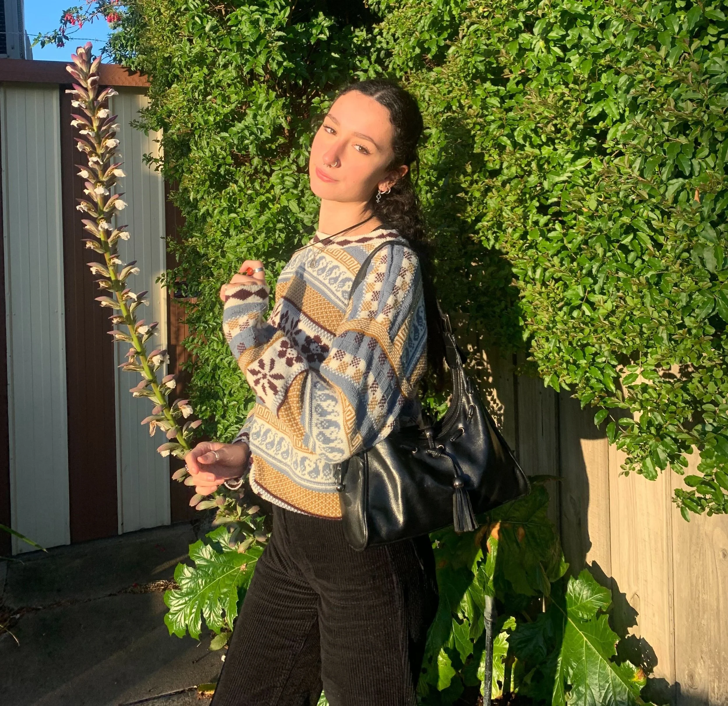 A young woman with dark curly hair, wearing a patterned sweater and black pants, standing outdoors in front of green foliage and a wooden fence, holding a small plant branch.