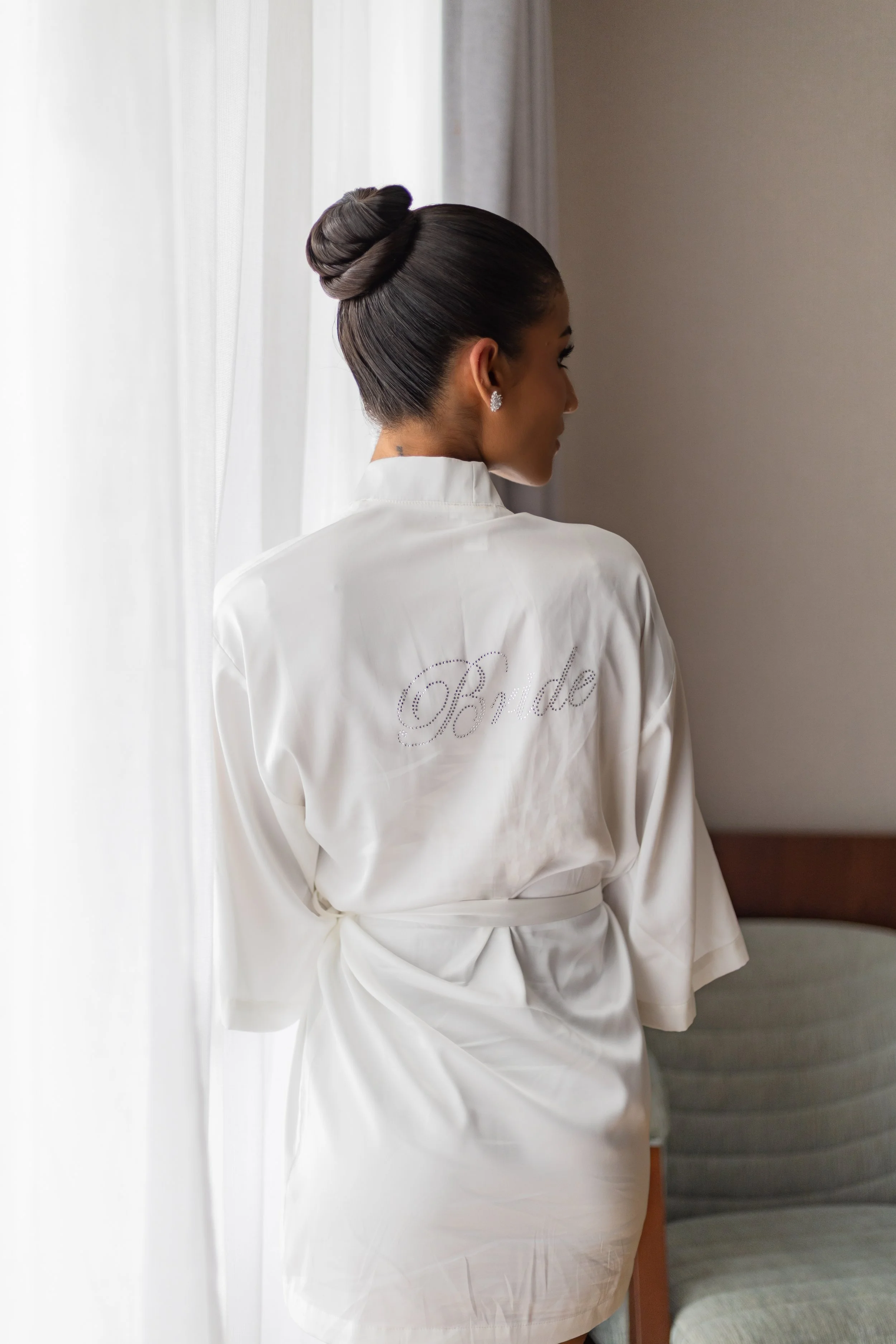 A woman with her hair in a bun, wearing a white satin robe with 'Bride' written on the back, standing near a window with sheer curtains.
