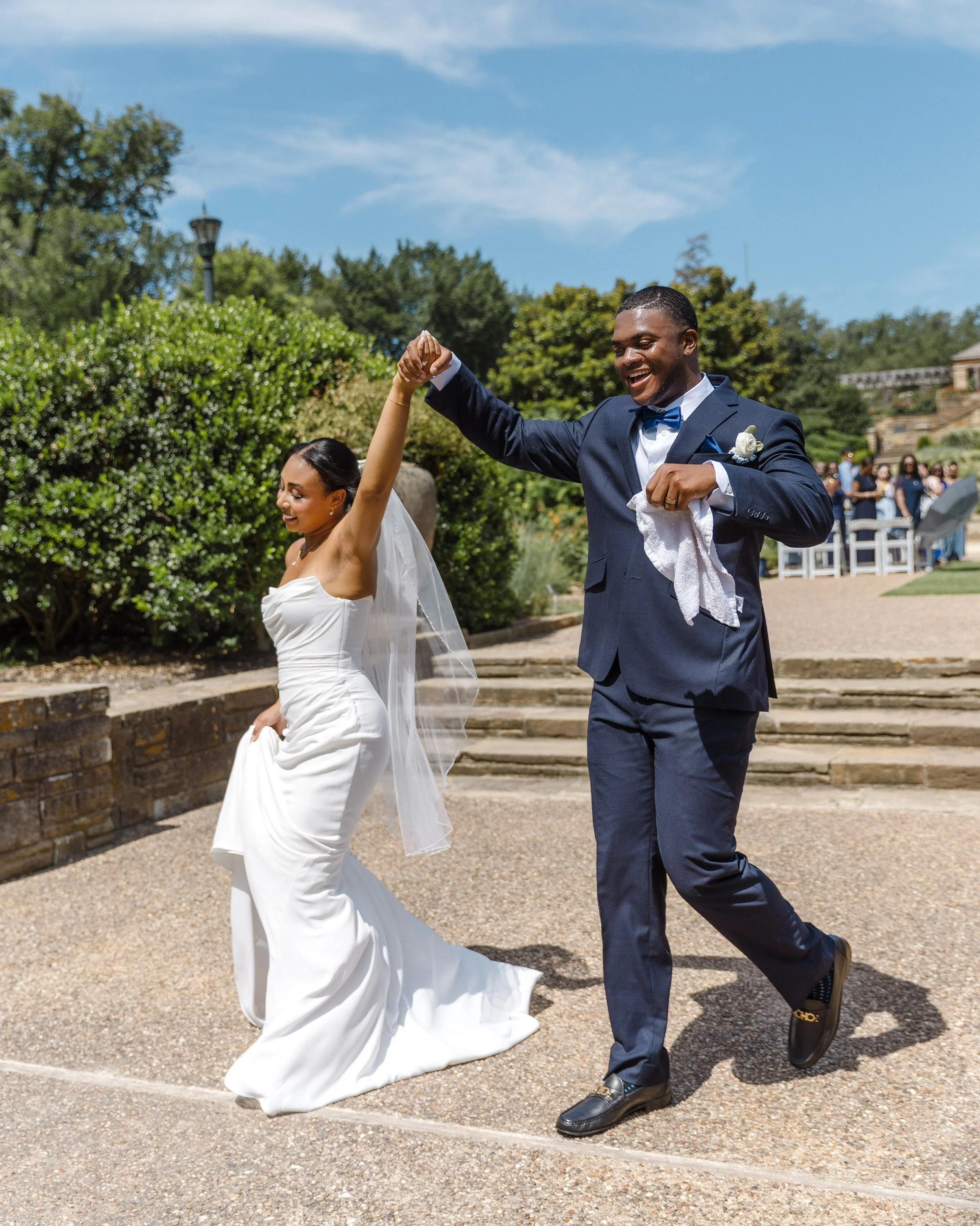 A newlywed couple dancing outdoors during their wedding celebration, with the bride in a white gown and the groom in a navy suit, smiling and holding hands.