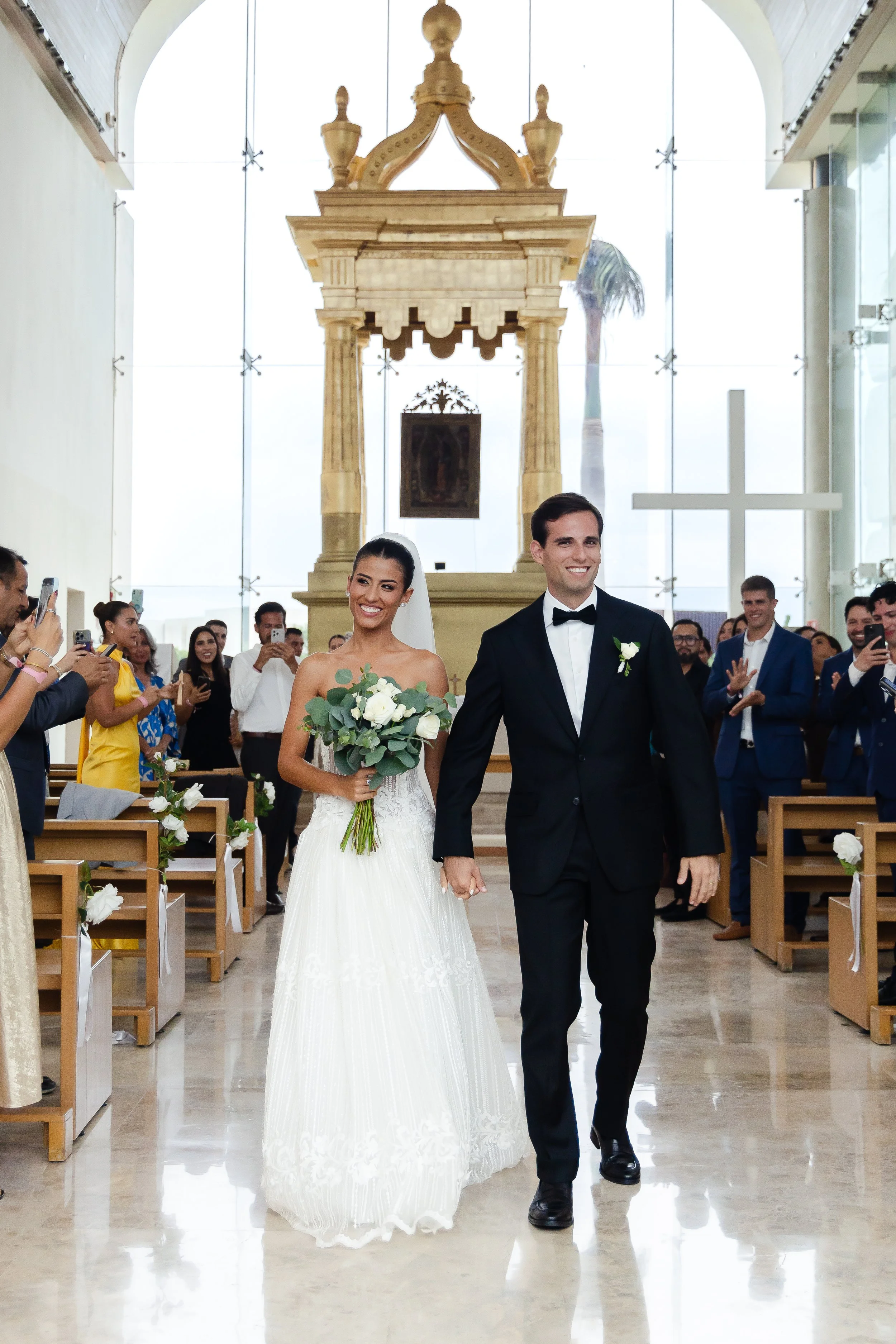 Bride and groom walking down the aisle of a church after their wedding ceremony, with guests taking photos and applauding.