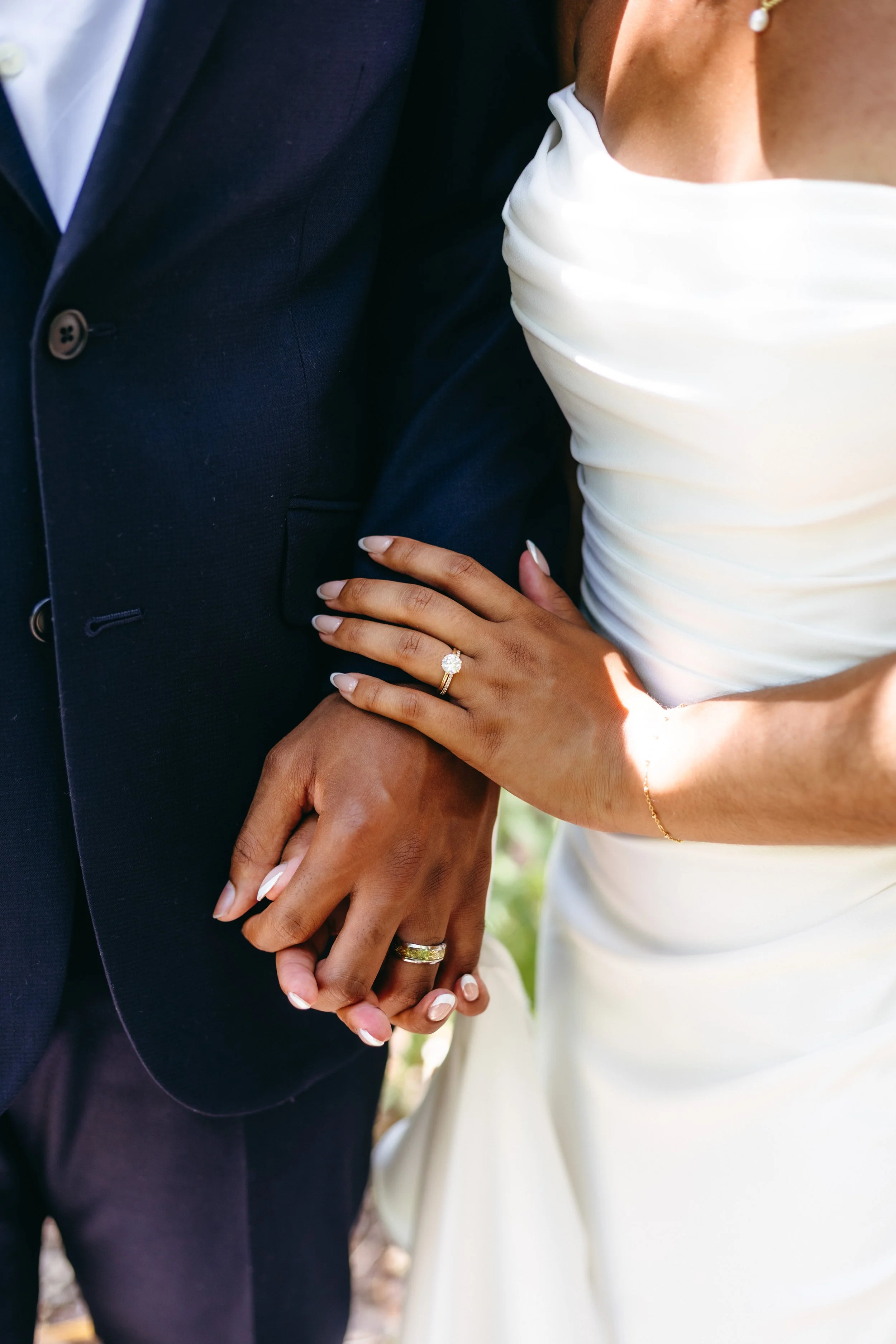 Close-up of a bride and groom holding hands, showing wedding rings, with the bride's hand resting on the groom's arm.