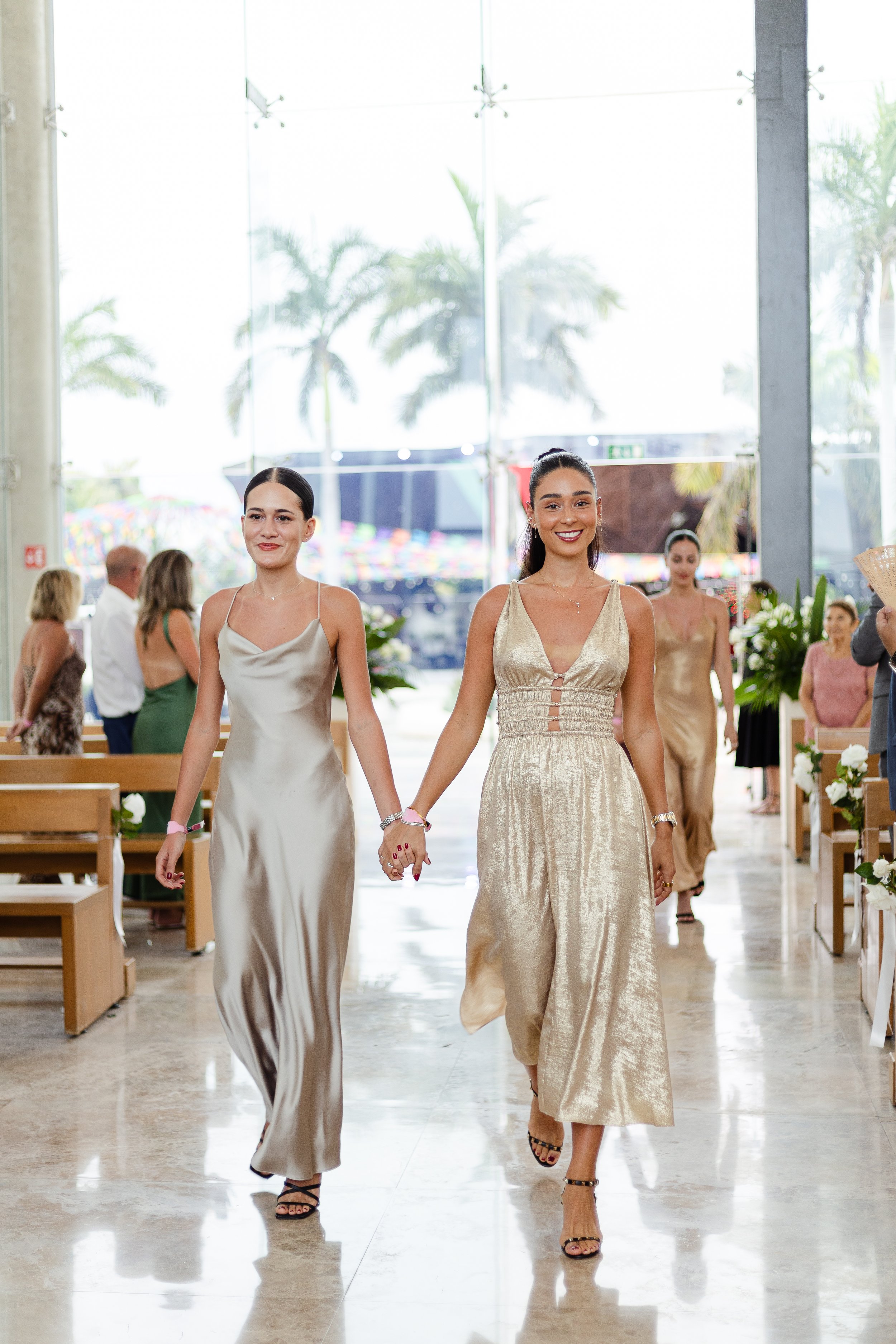 Two women in elegant dresses walk hand in hand down the aisle at a wedding, with other guests sitting and standing on either side and large windows showing palm trees outside.