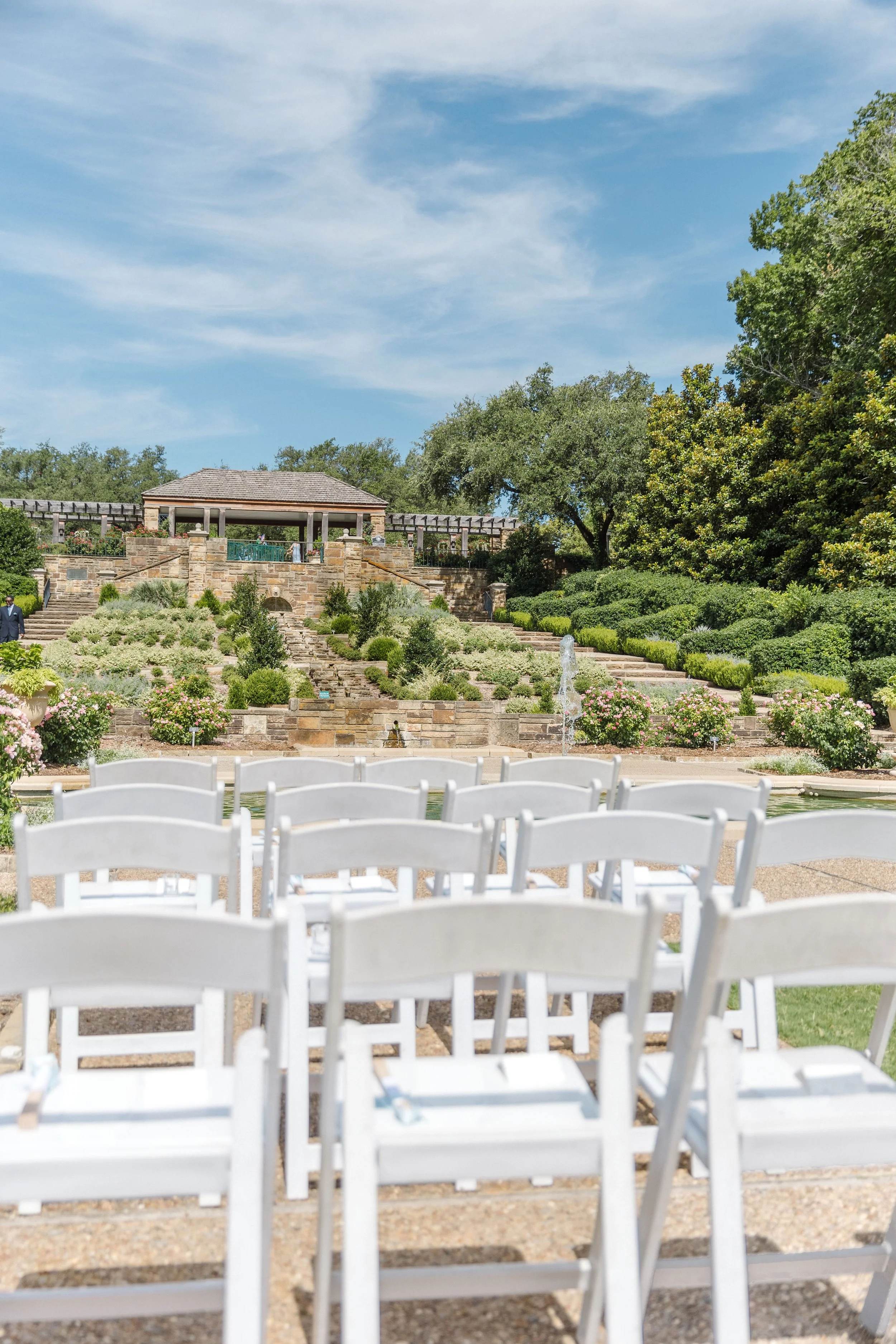 Outdoor wedding or event setup with white chairs arranged facing a garden with a tiered waterfall and fountain, lush greenery, flowering bushes, and a pavilion under a blue sky with scattered clouds.