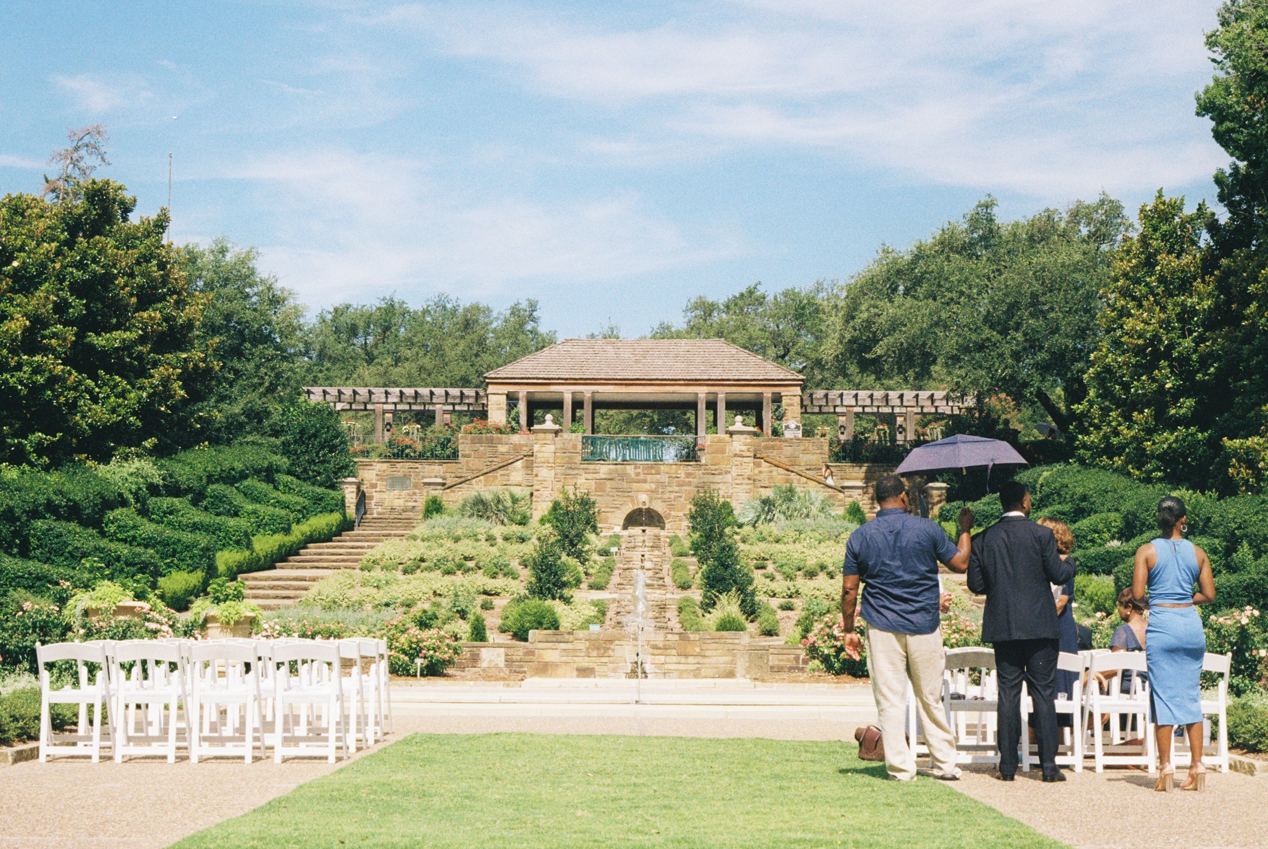 People setting up chairs for an outdoor wedding ceremony in a lush garden with a tiered stone fountain and pavilion in the background.