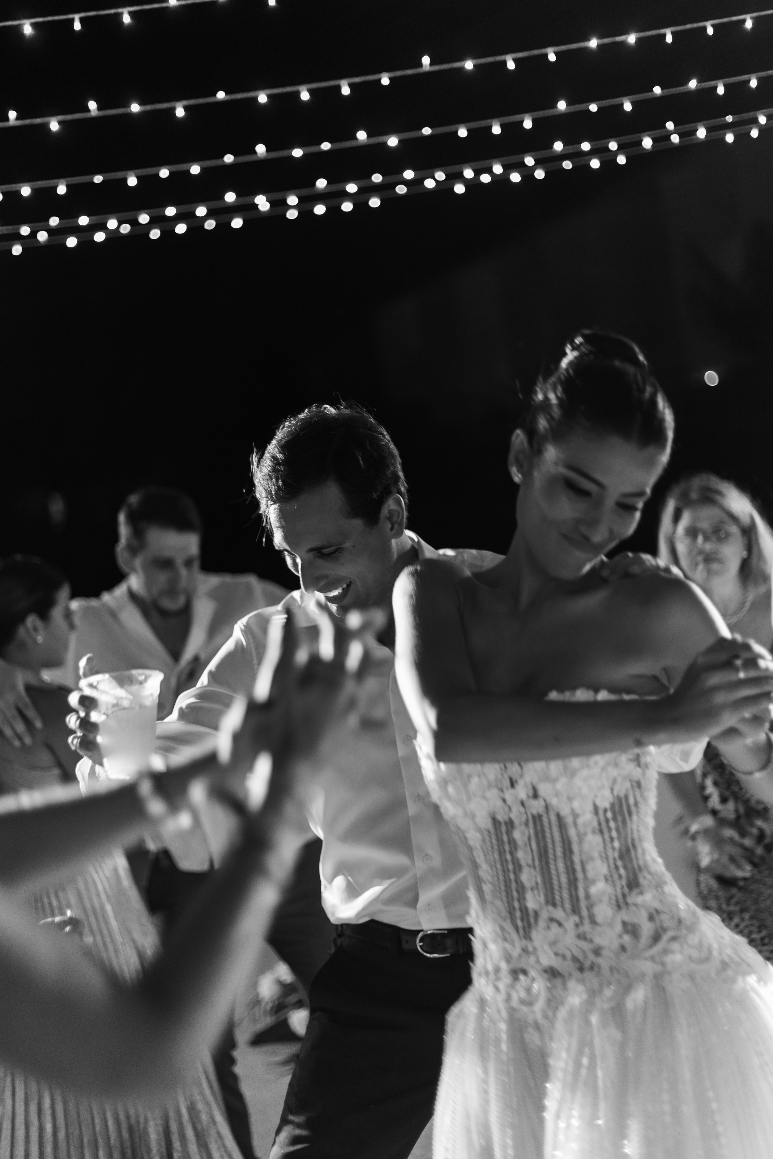 People dancing at a celebration, with a couple in the foreground dancing closely, smiling, and others in the background enjoying the event under string lights at night.