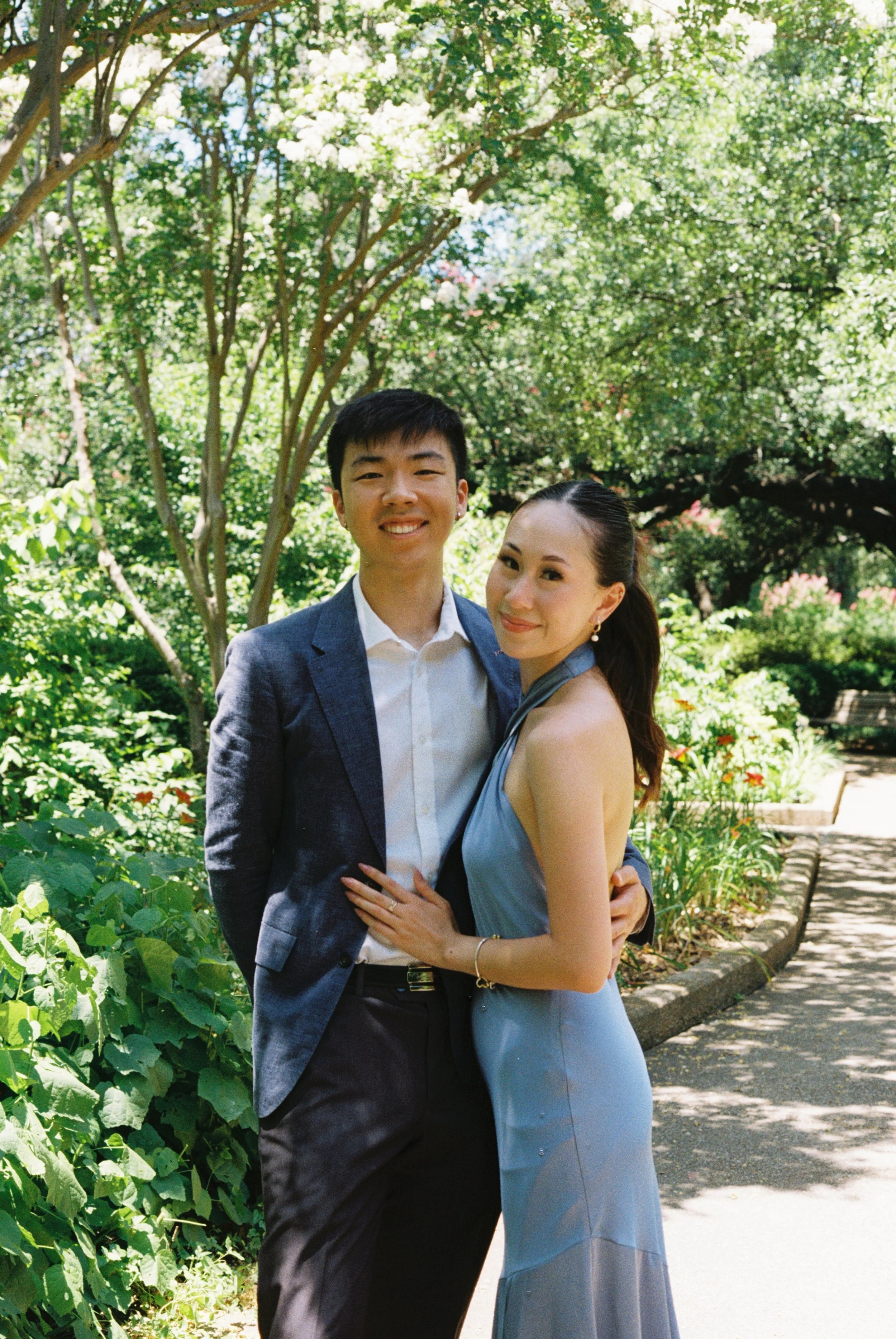 A couple standing outside in a park, smiling at the camera. The man is wearing a suit jacket and white shirt, and the woman is wearing a light blue dress.