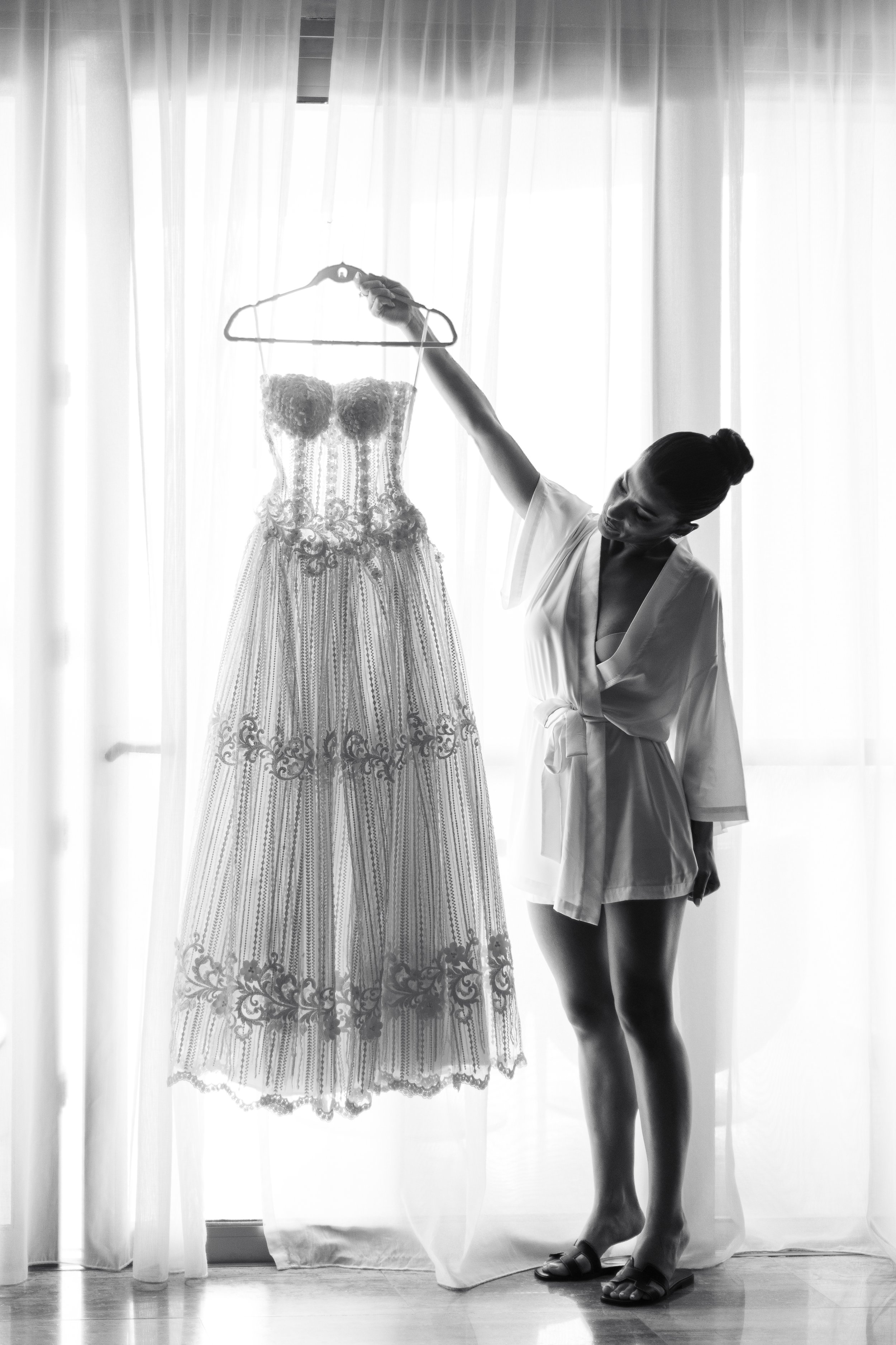 A woman in a dressing gown holds up a wedding dress in front of sheer curtains with sunlight streaming in.