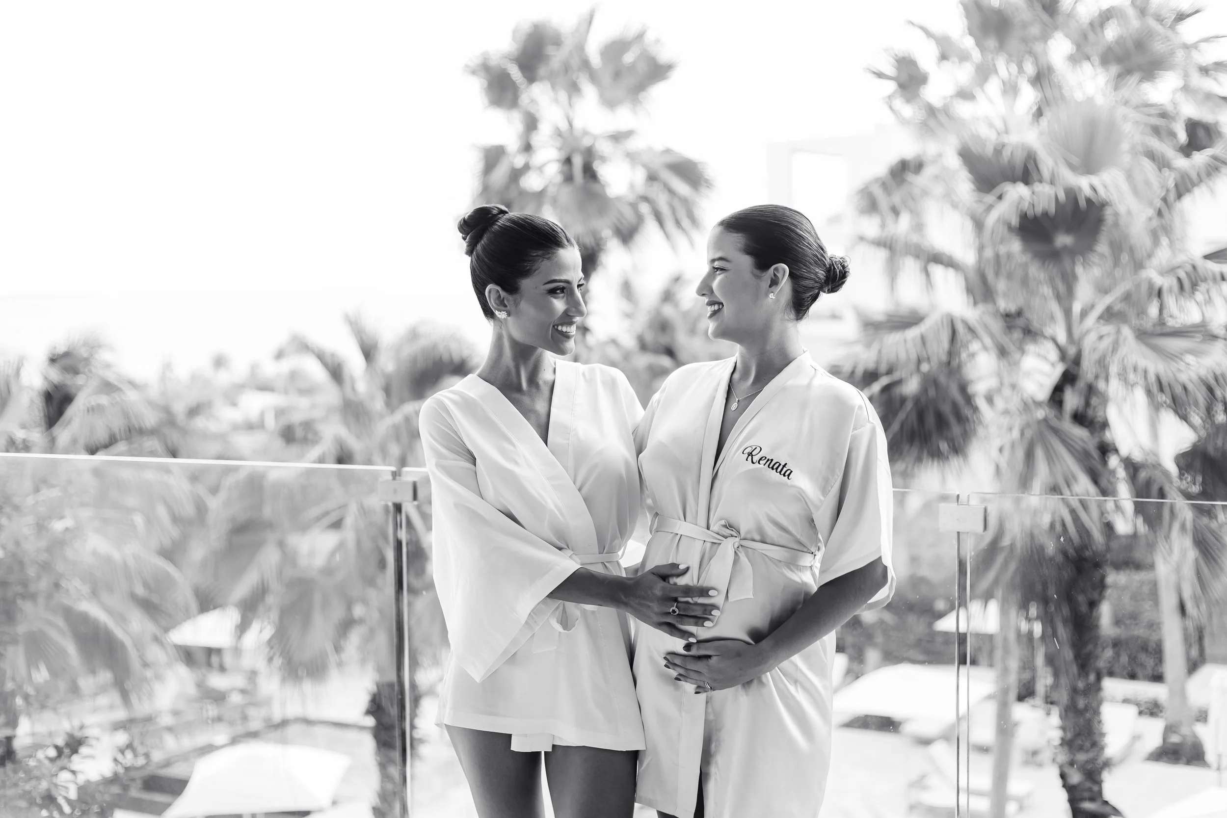 Two women wearing robes, smiling and holding their pregnant bellies, standing outdoors with palm trees in the background.