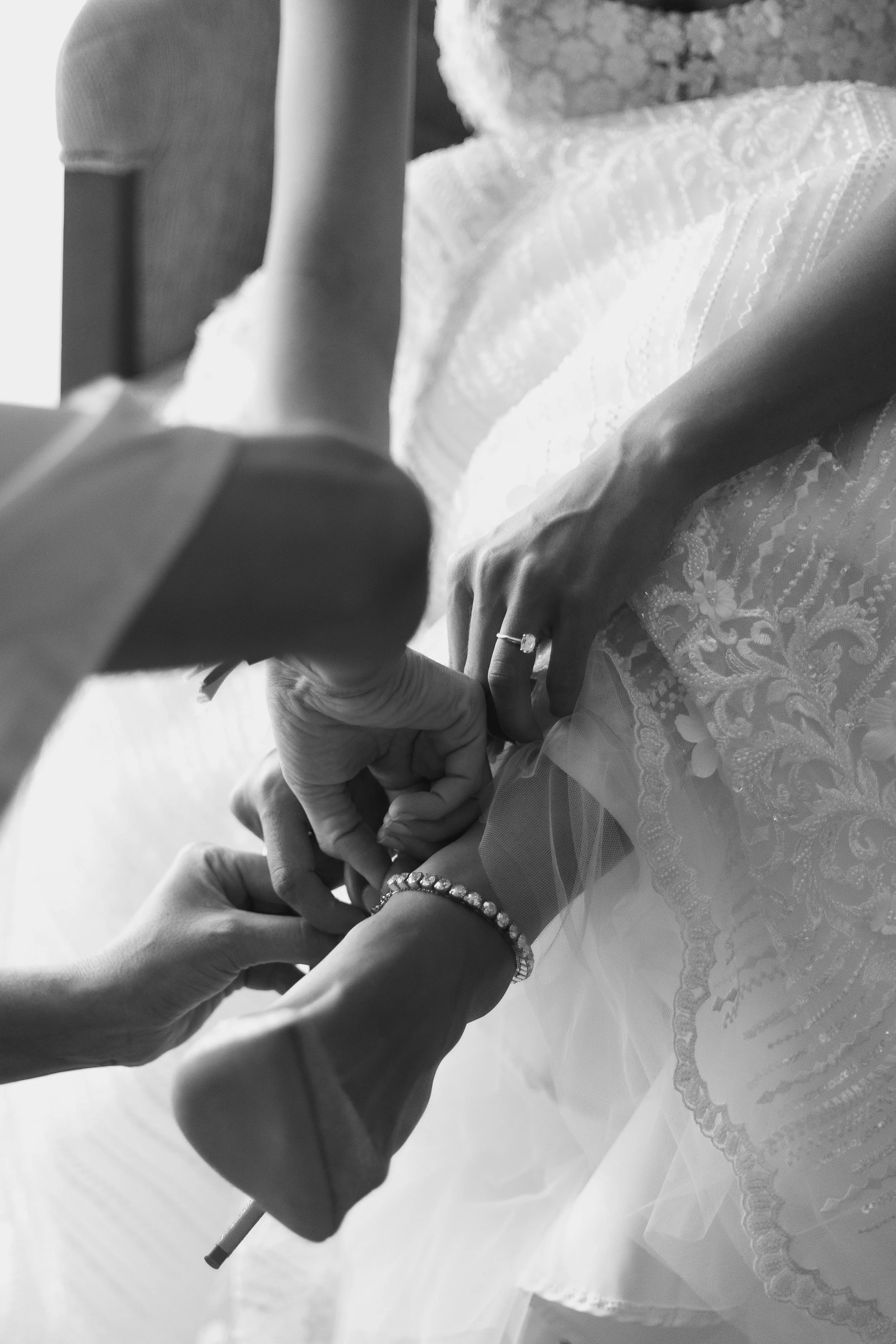Close-up of a bride getting ready with assistance from others, focusing on her hands and jewelry, on her wedding dress in black and white.