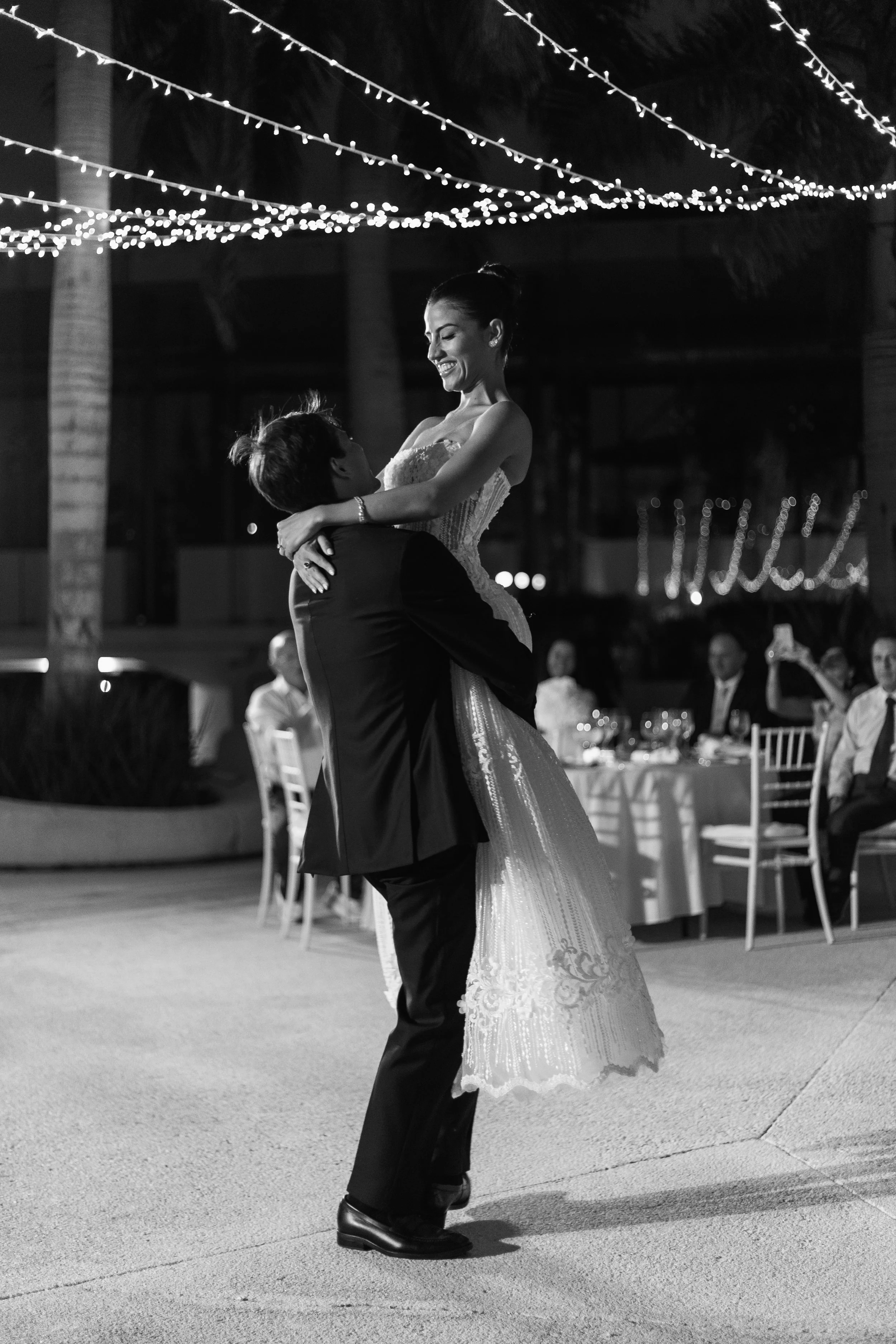 A newlywed couple is dancing at their wedding reception, with the groom lifting the bride into the air. The scene is illuminated by string lights overhead, and guests are seated at tables in the background.