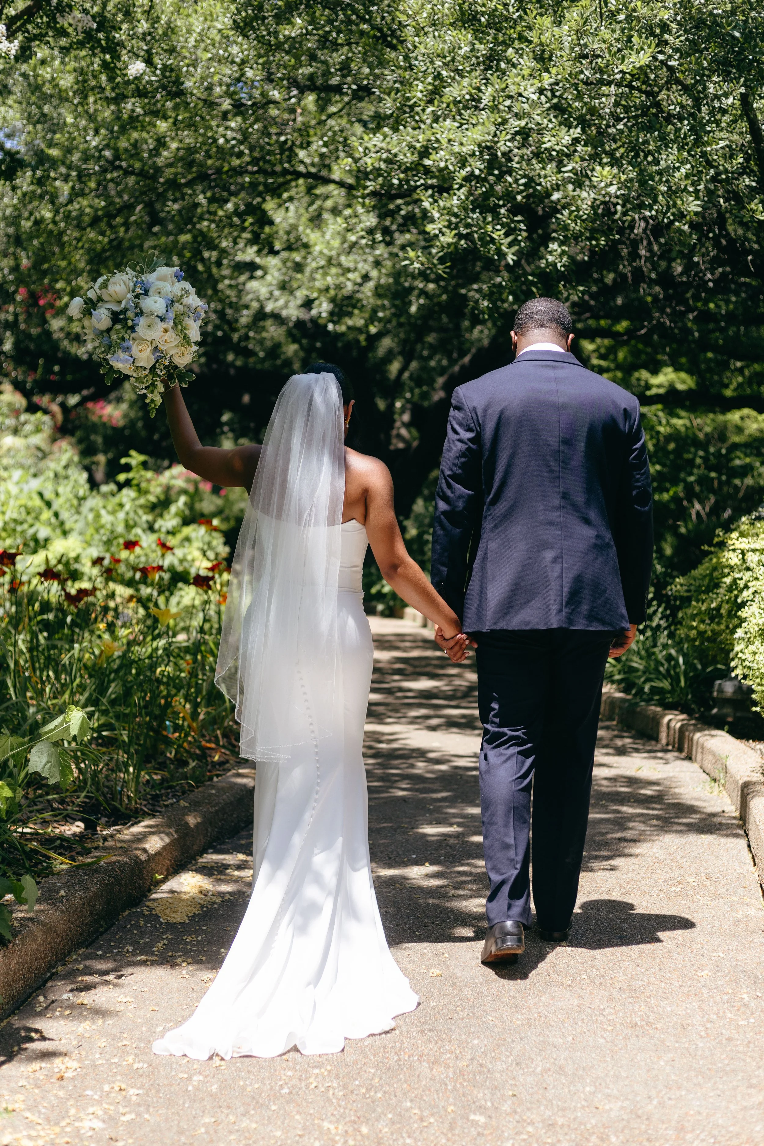 Bride and groom walking hand in hand on a garden pathway, with the bride holding a bouquet of flowers in the air.