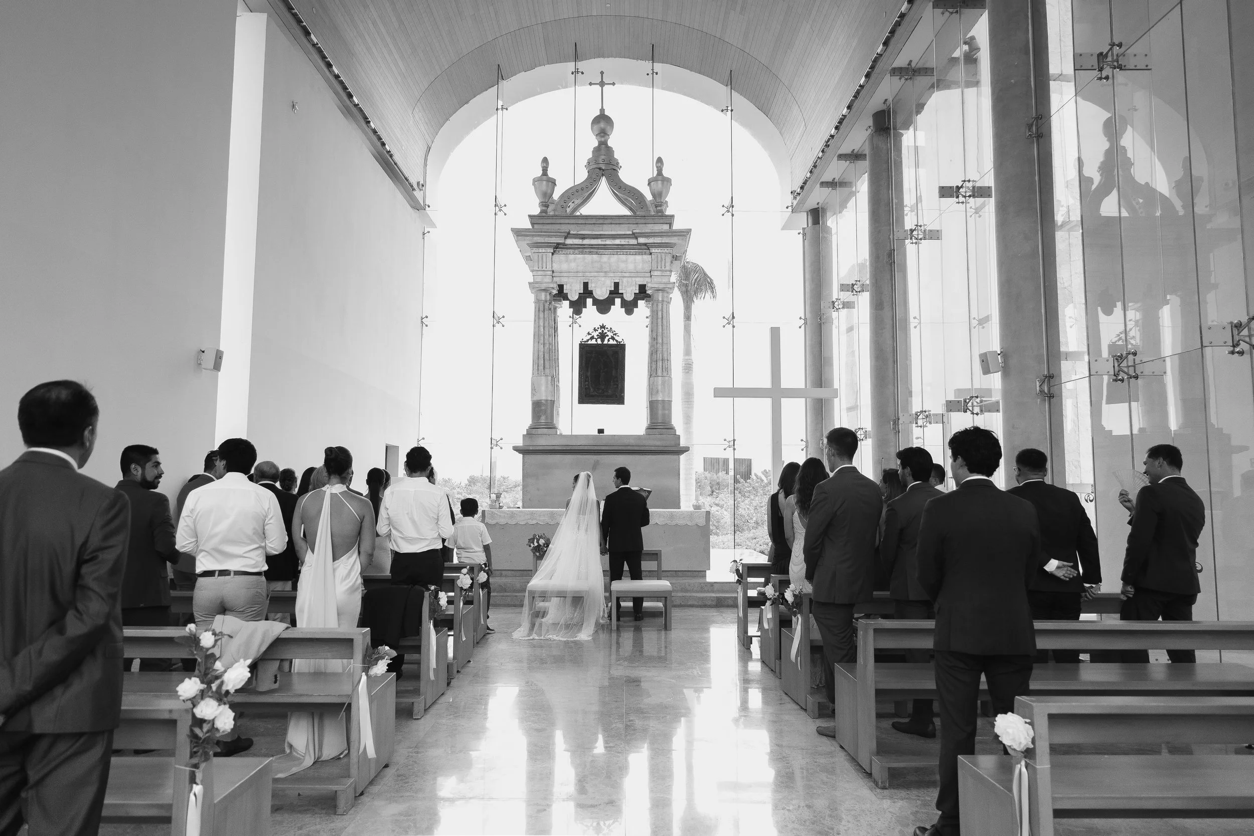 A black and white photo of a wedding ceremony inside a modern church with tall glass windows. The bride and groom stand facing the altar with guests on either side, and a large cross is visible at the front.