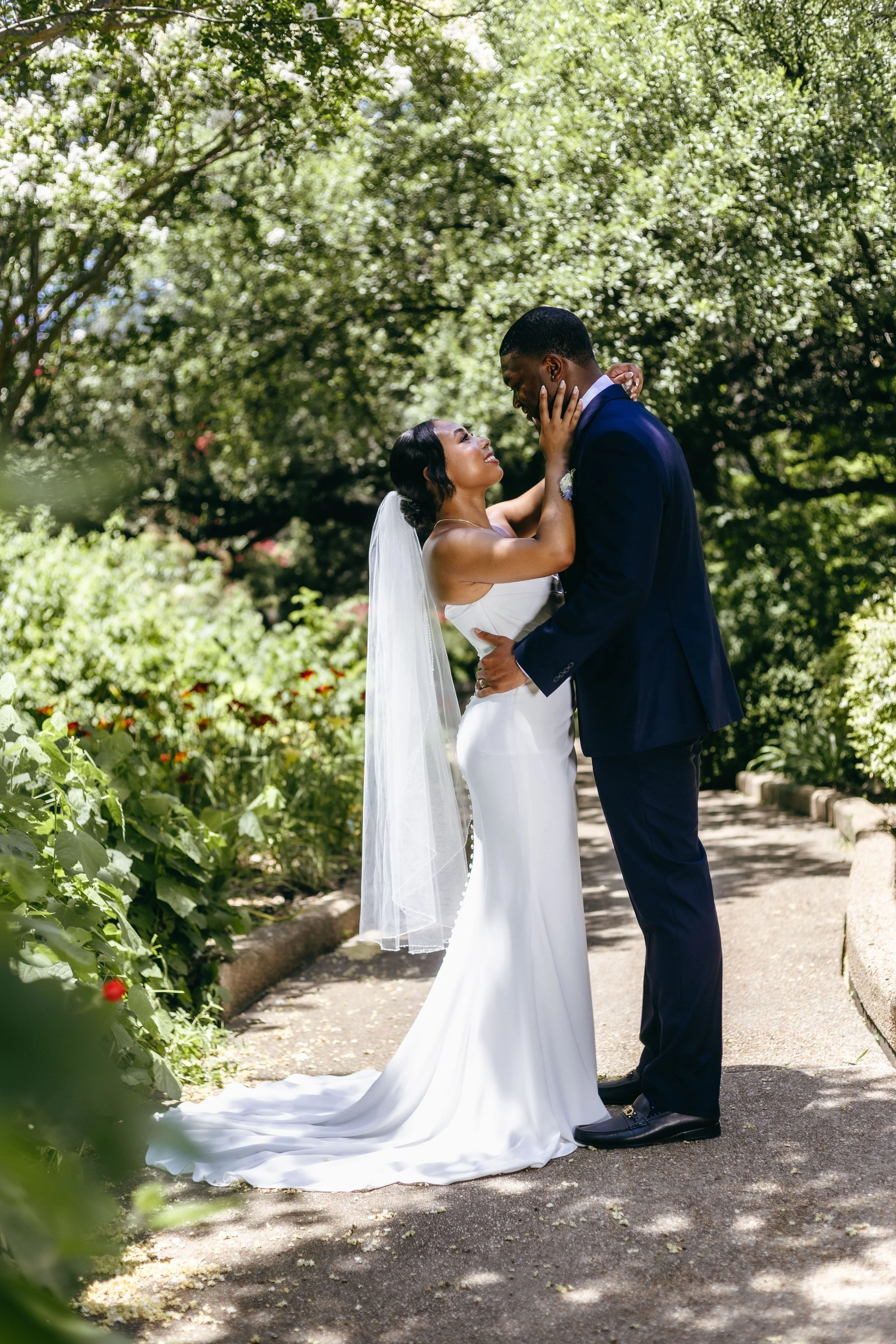 A bride and groom stand close together on a garden path, embracing and gazing into each other's eyes. The bride is wearing a white wedding gown with a veil, and the groom is dressed in a dark blue suit. They are surrounded by lush green trees and pla