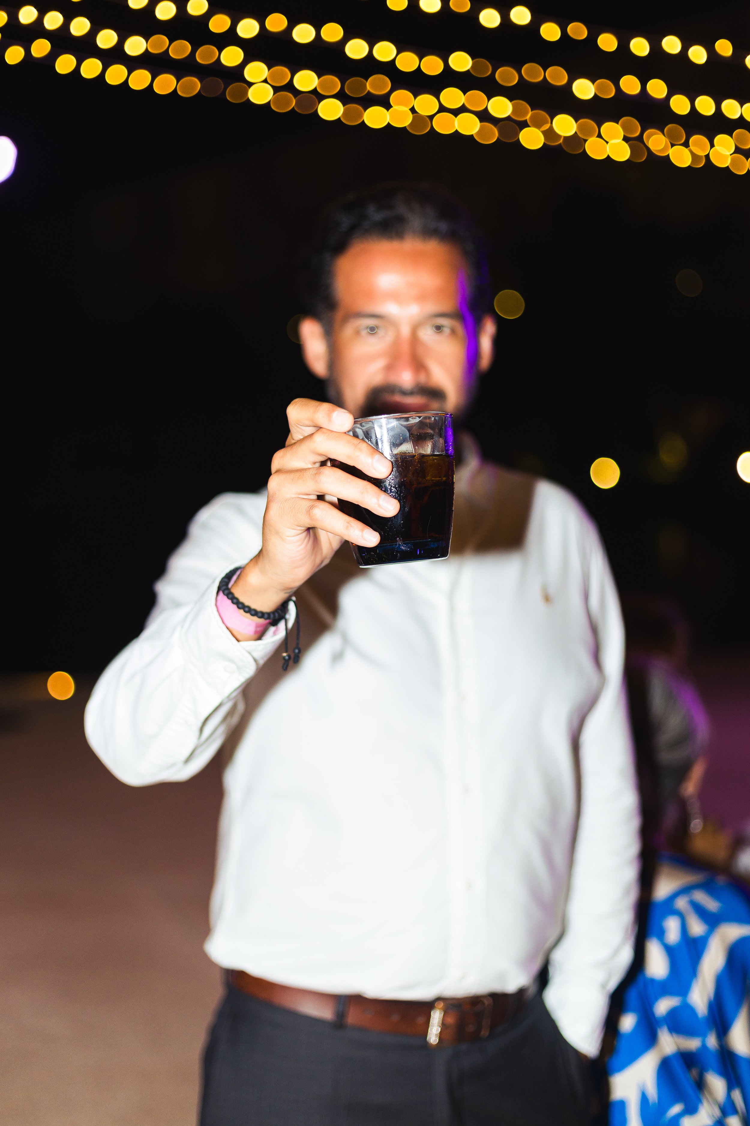 A man in a white shirt holding a glass of dark soda at night, with blurred string lights in the background.