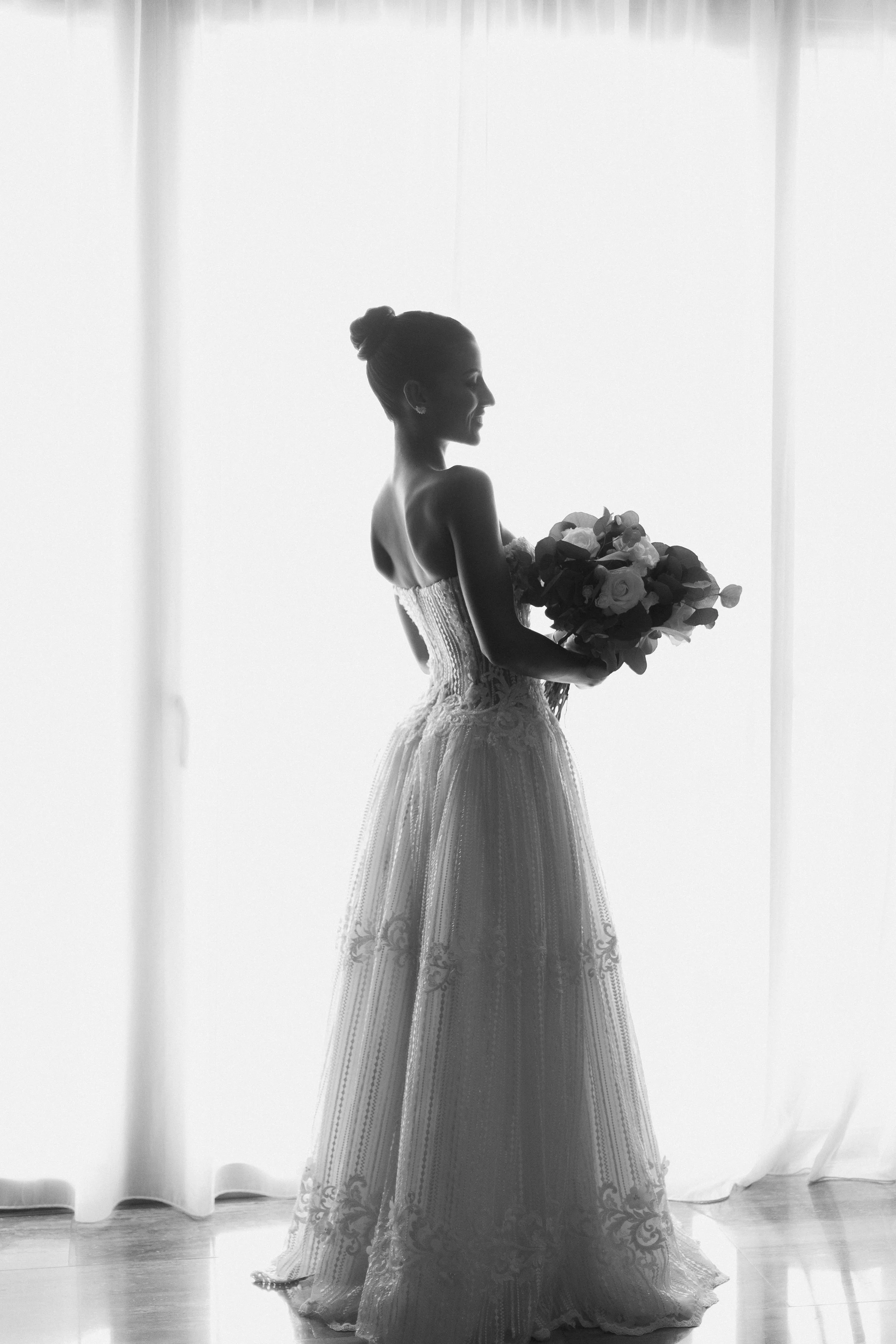 Silhouette of a woman in a wedding dress holding a bouquet of flowers in front of a sheer white curtain.