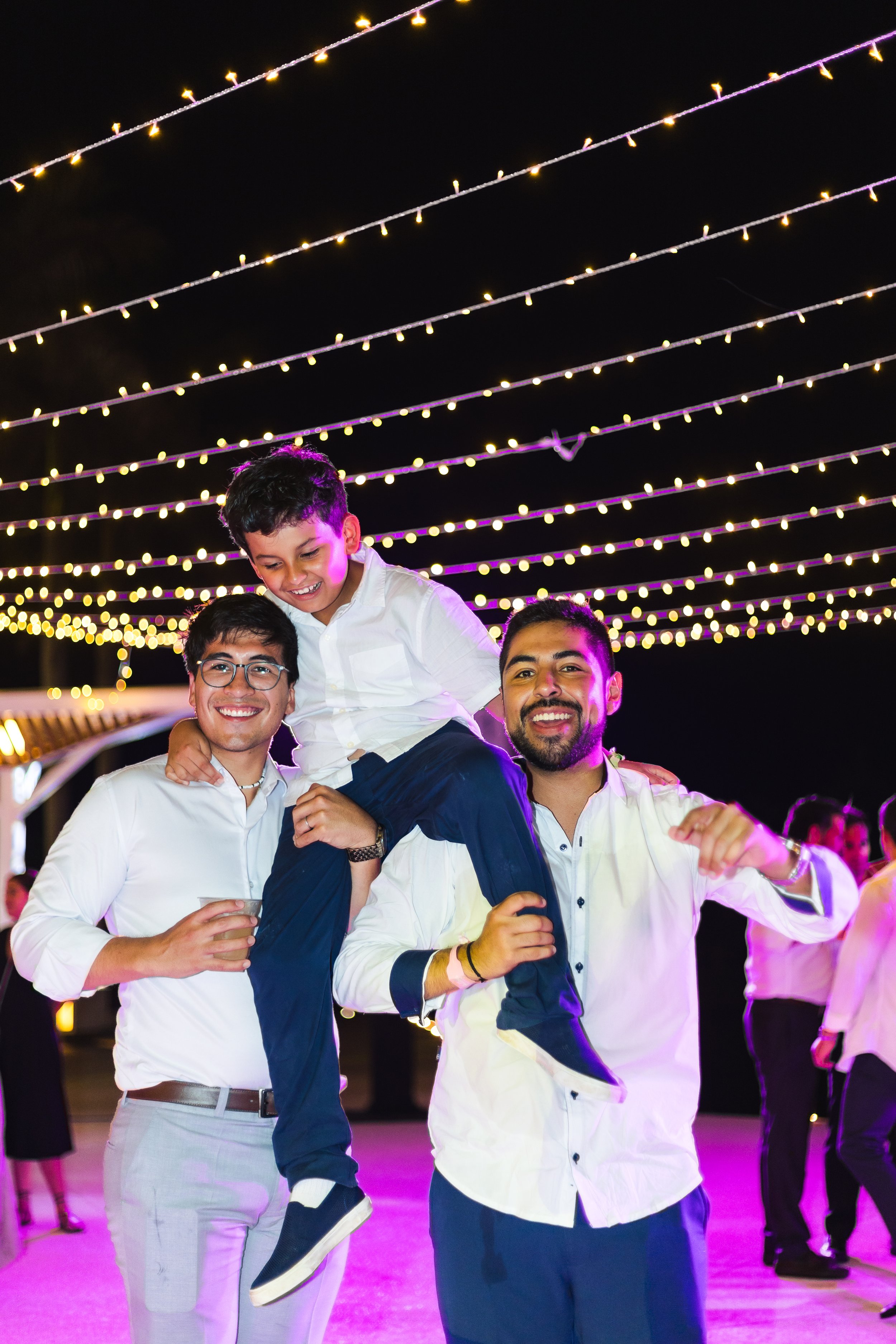 Three men and a boy celebrating at night under string lights, with one man carrying the boy on his shoulders, all smiling.