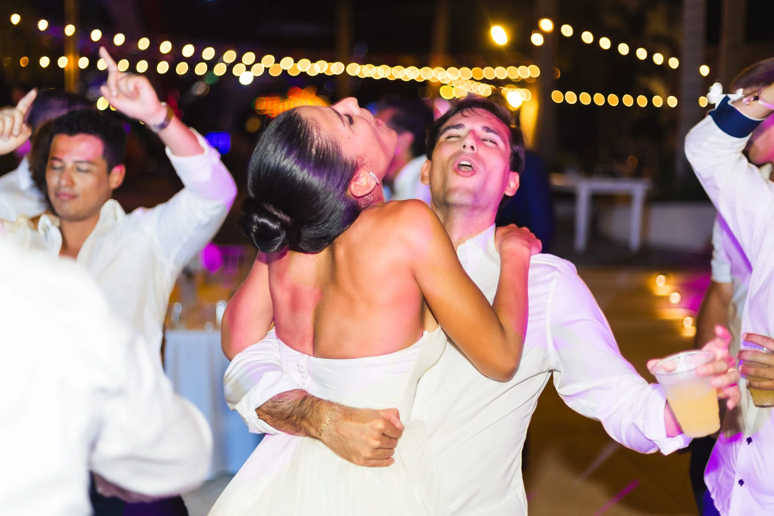 People dancing and celebrating at a lively outdoor party with string lights in the background.