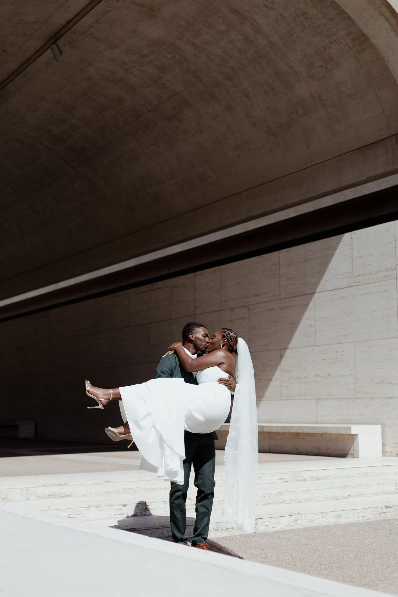 A groom holding a bride in a white wedding dress under a modern concrete archway.