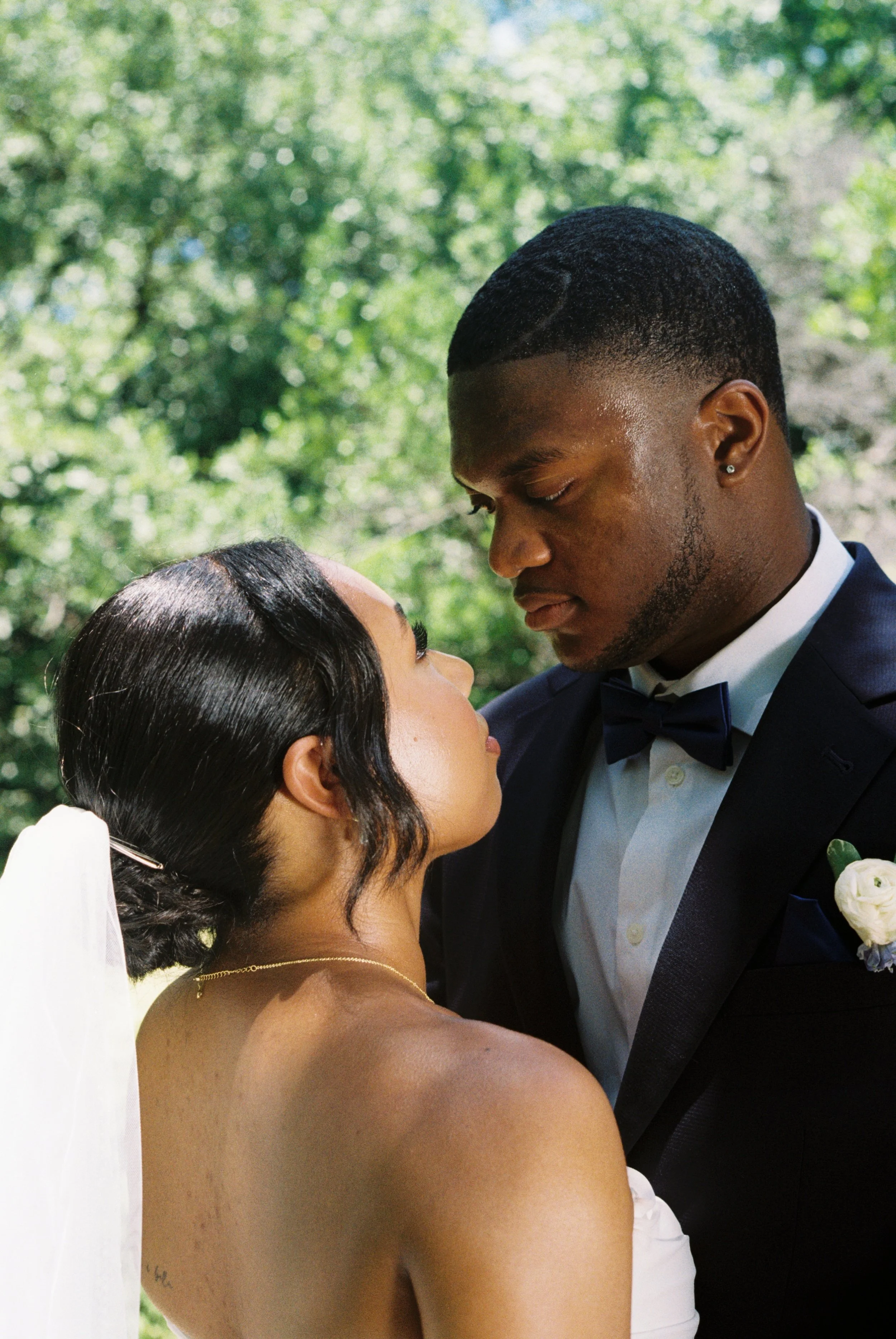 A bride and groom stand close together outdoors, gazing into each other's eyes, with lush green trees in the background.