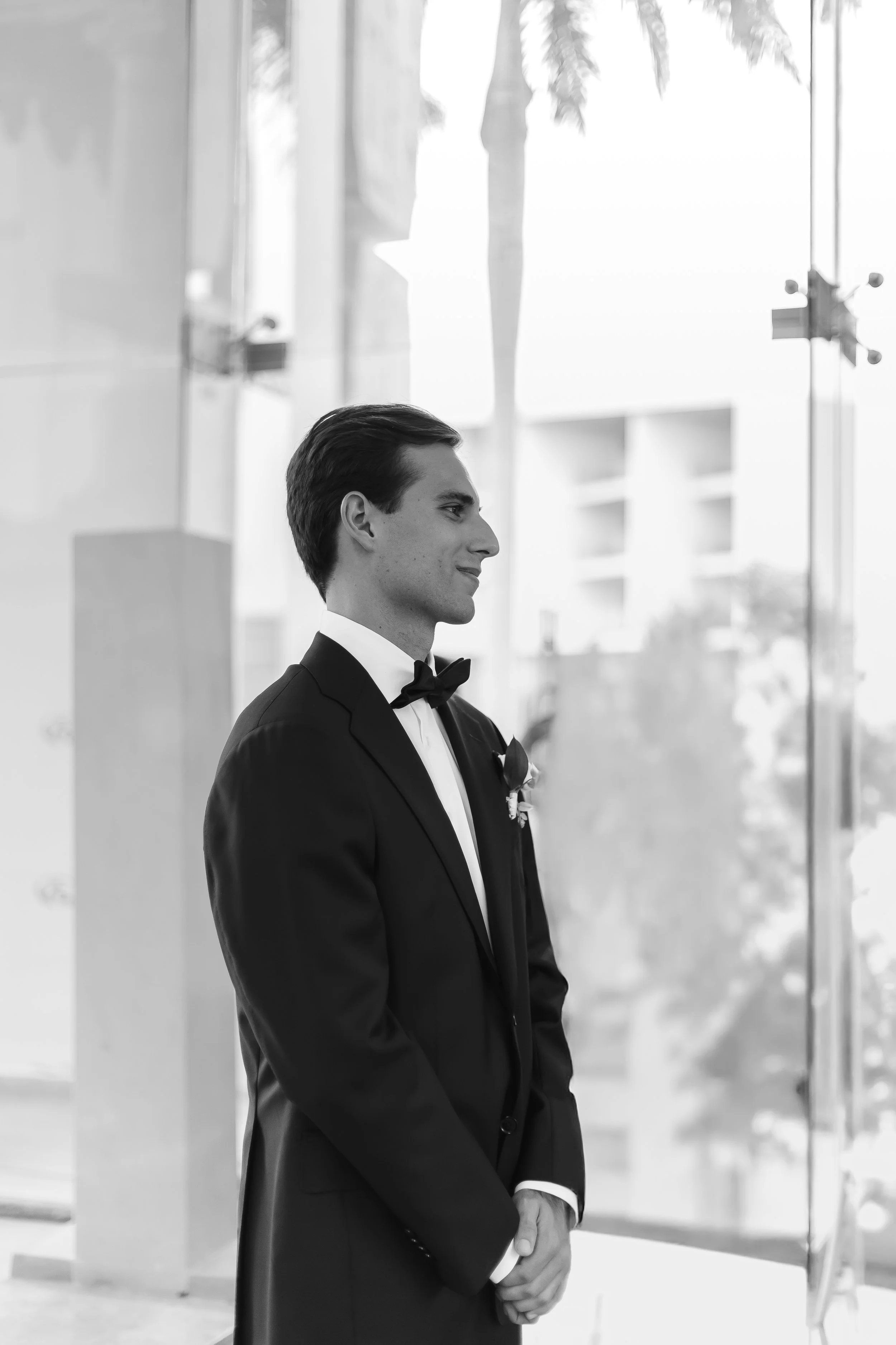 A man in a tuxedo standing indoors, smiling, with large windows behind him showing trees and buildings.