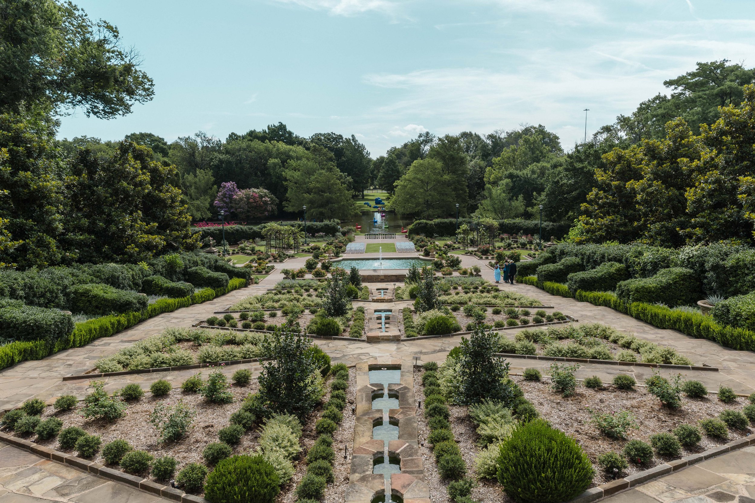 A view of a manicured garden with unpaved pathways, symmetrical plant beds with small bushes and flowers, and a water feature in the distance. Tall trees surround the garden under a partly cloudy sky.