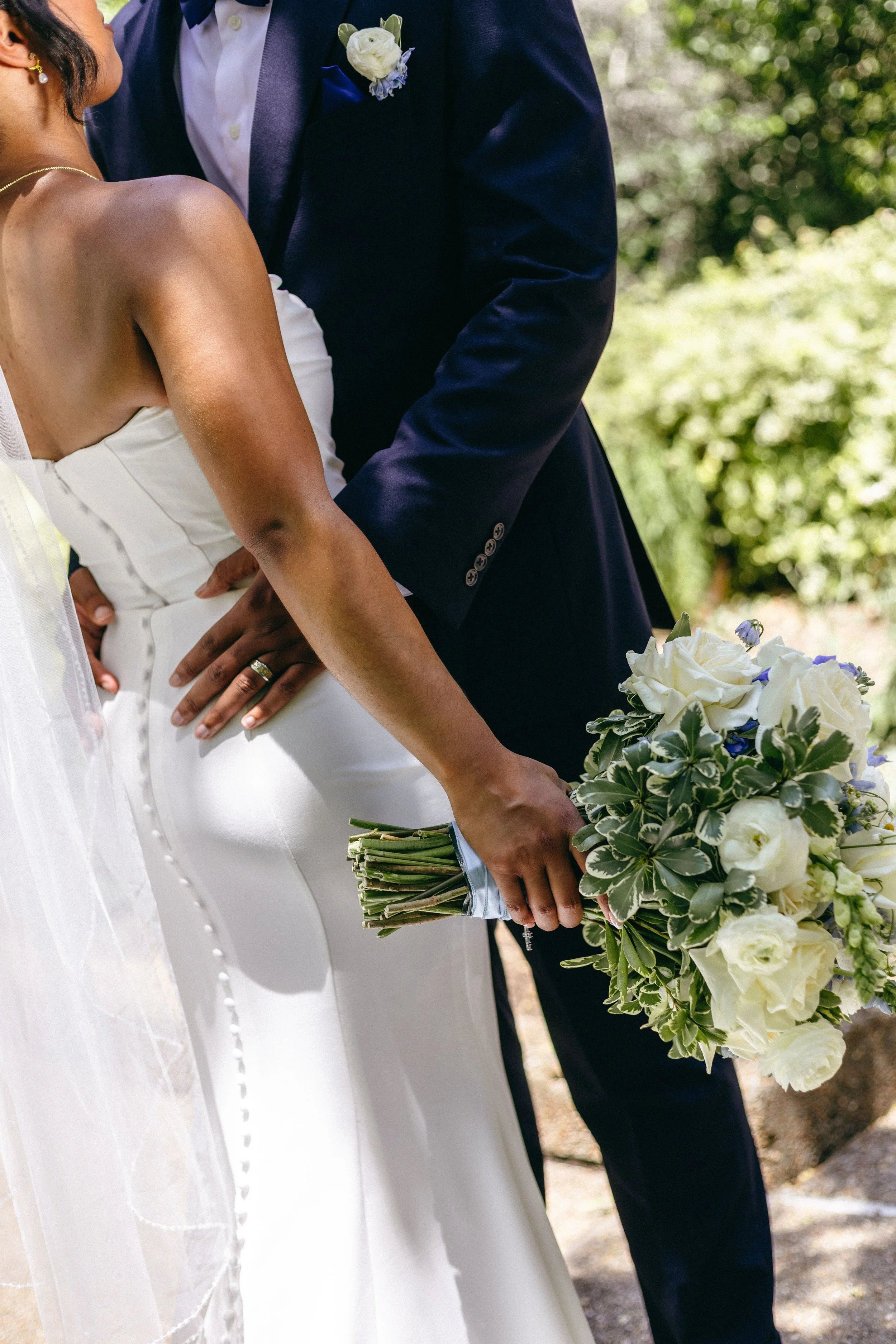 Close-up of a bride and groom standing outdoors, the bride holding a bouquet of white and purple flowers, the groom wearing a dark suit with a white shirt and boutonnière.