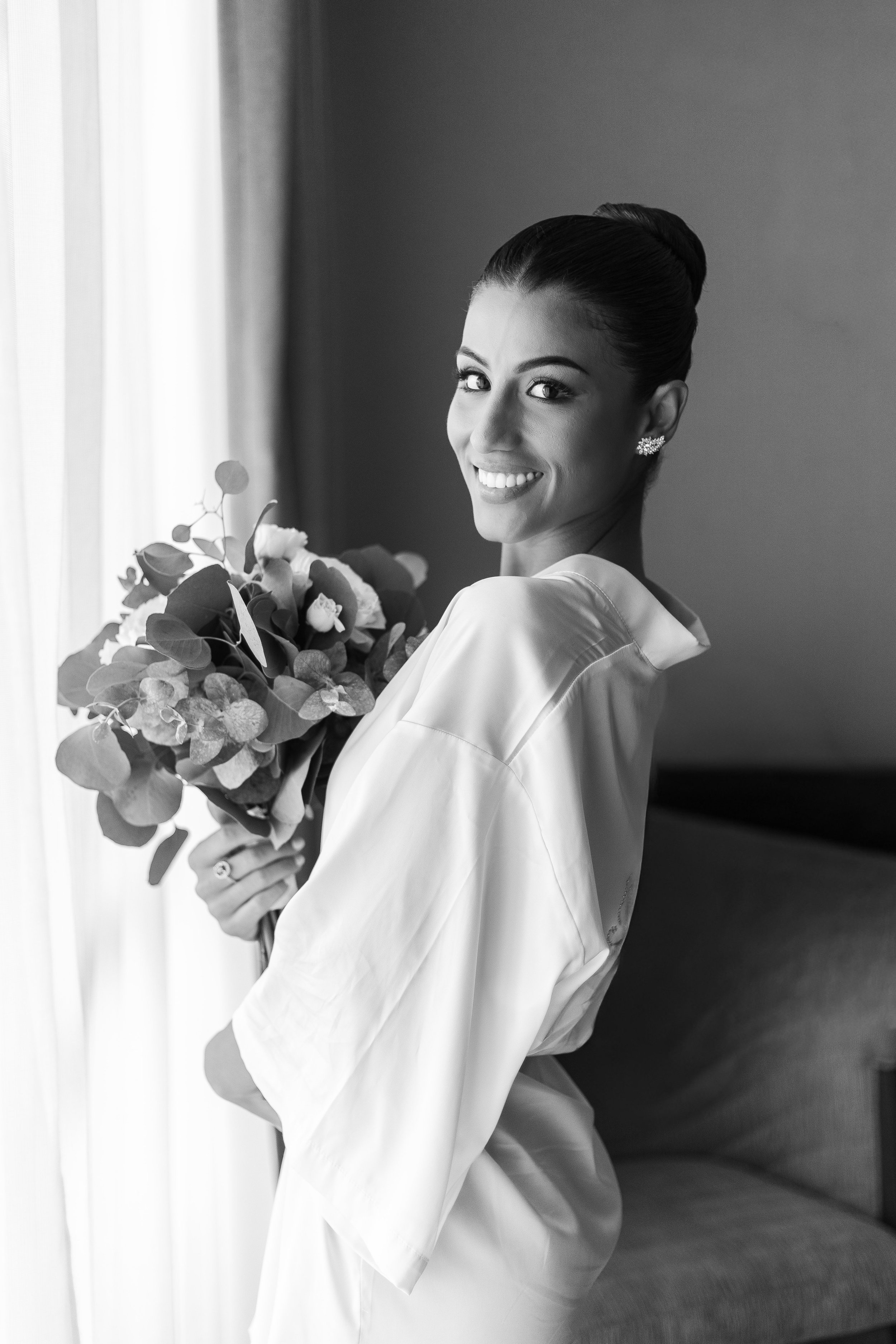 Black and white photo of a smiling bride holding a bouquet standing indoors near a window with curtains.