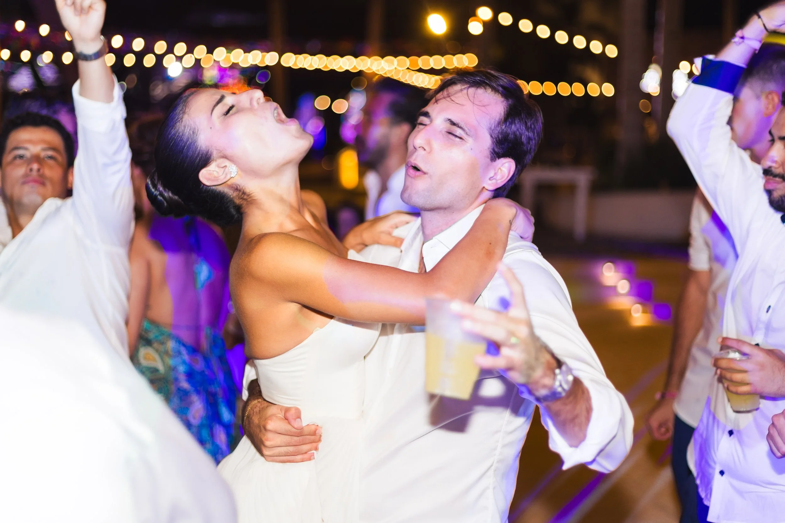 People dancing and celebrating at a lively party, with string lights in the background and drinks in hand.