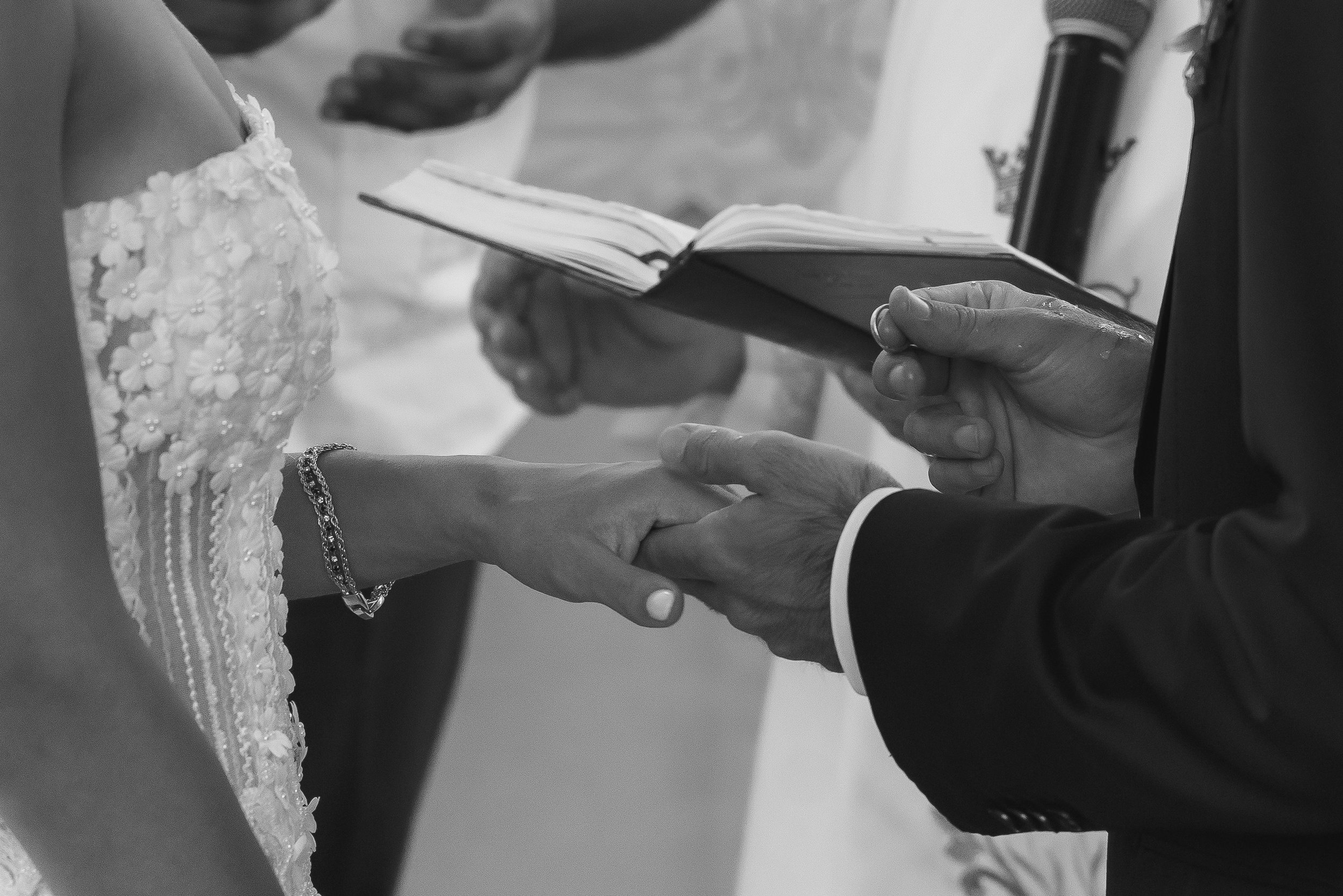 A bride and groom holding hands during their wedding ceremony, with the officiant reading from a book in the background.