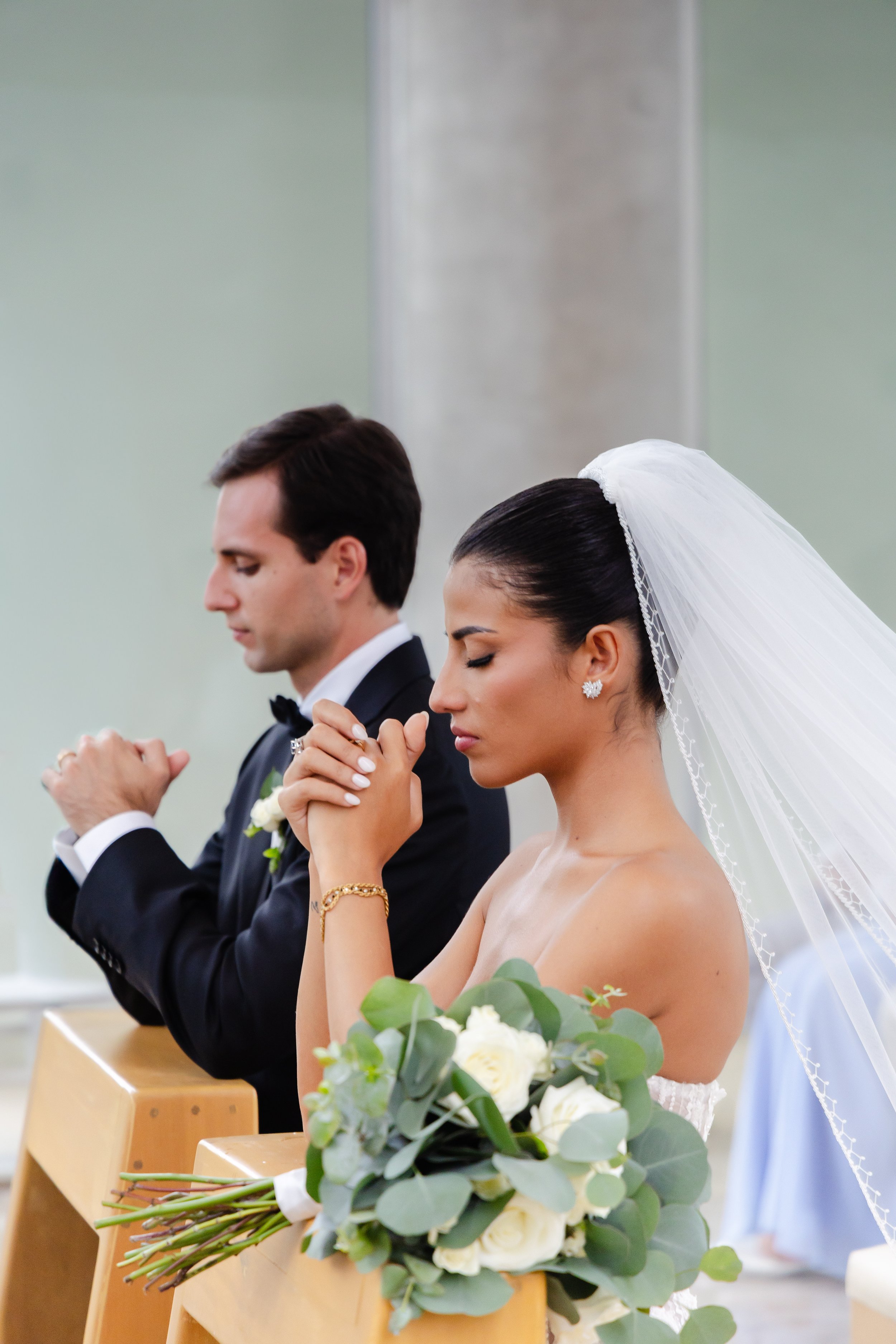 A bride and groom kneeling in prayer during a wedding ceremony with hands clasped and eyes closed, the bride holding a bouquet of white roses and green foliage, bride wearing a white veil and earrings, groom in a black suit and bow tie.