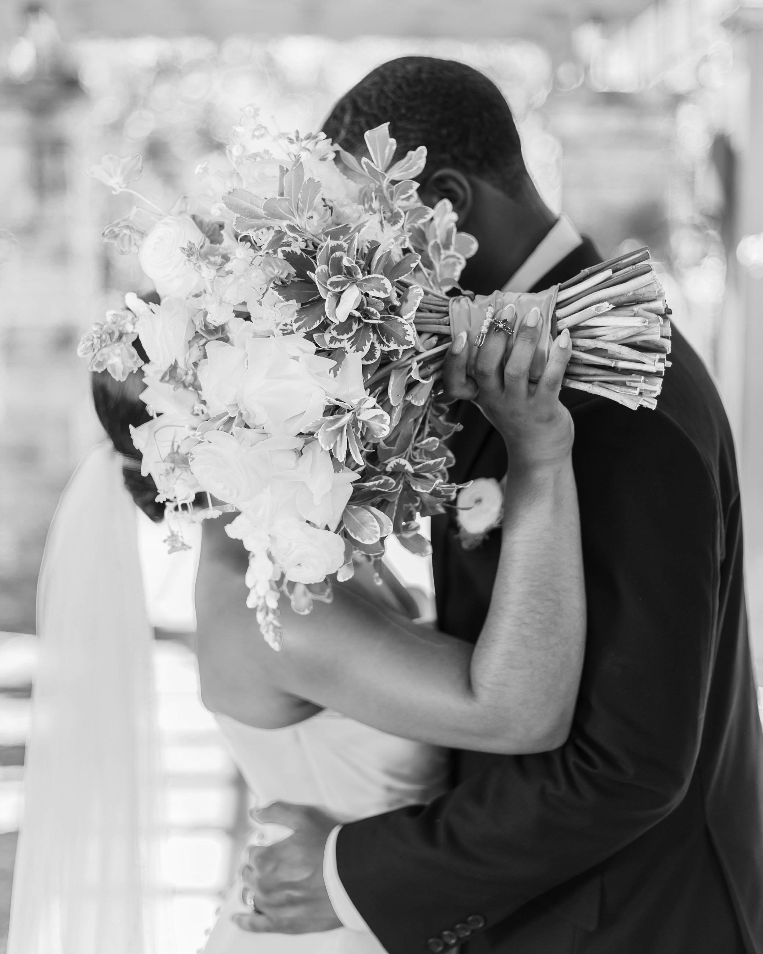A couple embraces, with the woman holding a large bouquet of flowers close to her face, partly hiding her face, on what appears to be a wedding day.