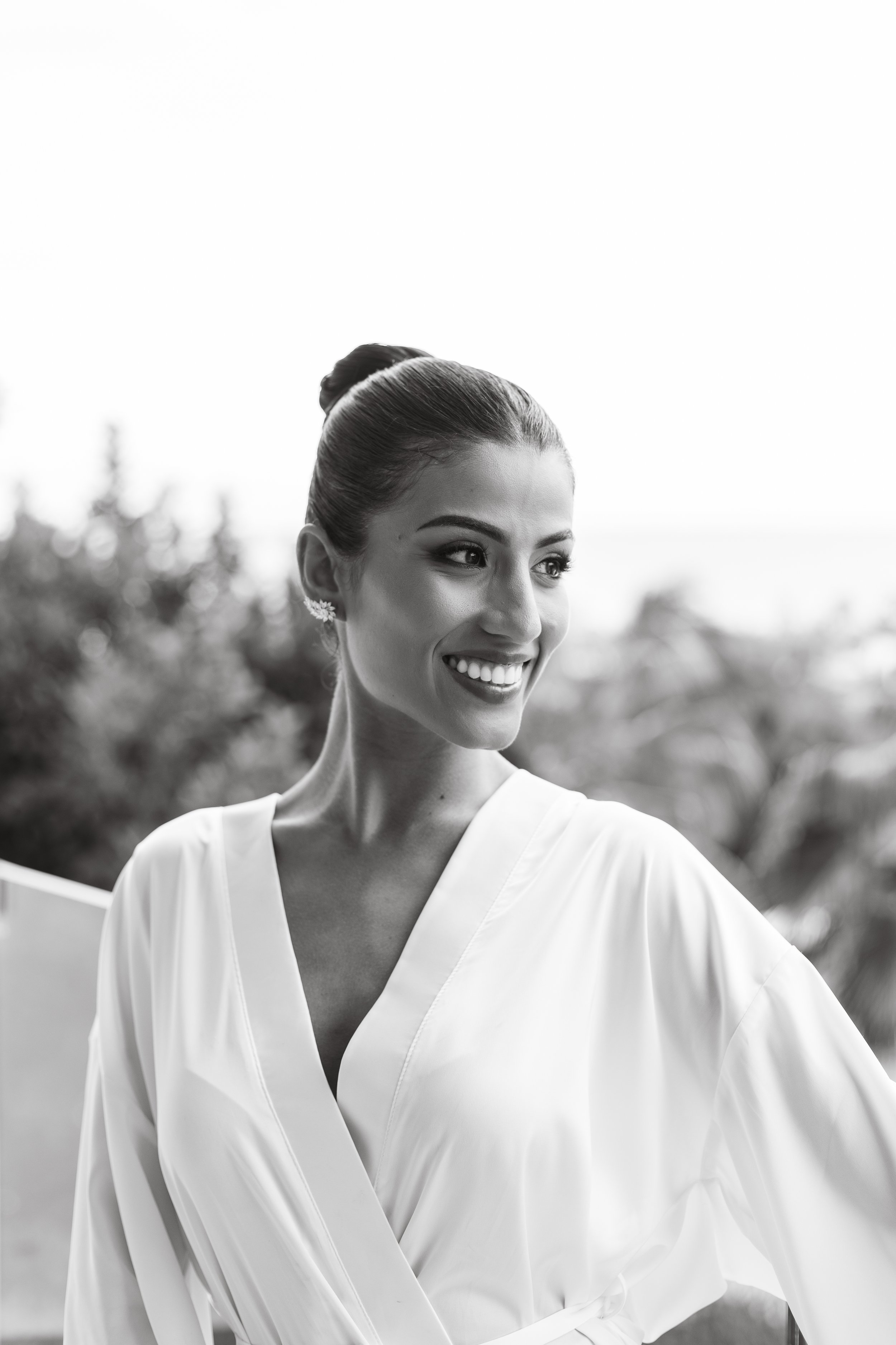 Black and white photo of a smiling woman with dark hair in a bun, wearing earrings and a white blouse, outdoors with trees in the background.