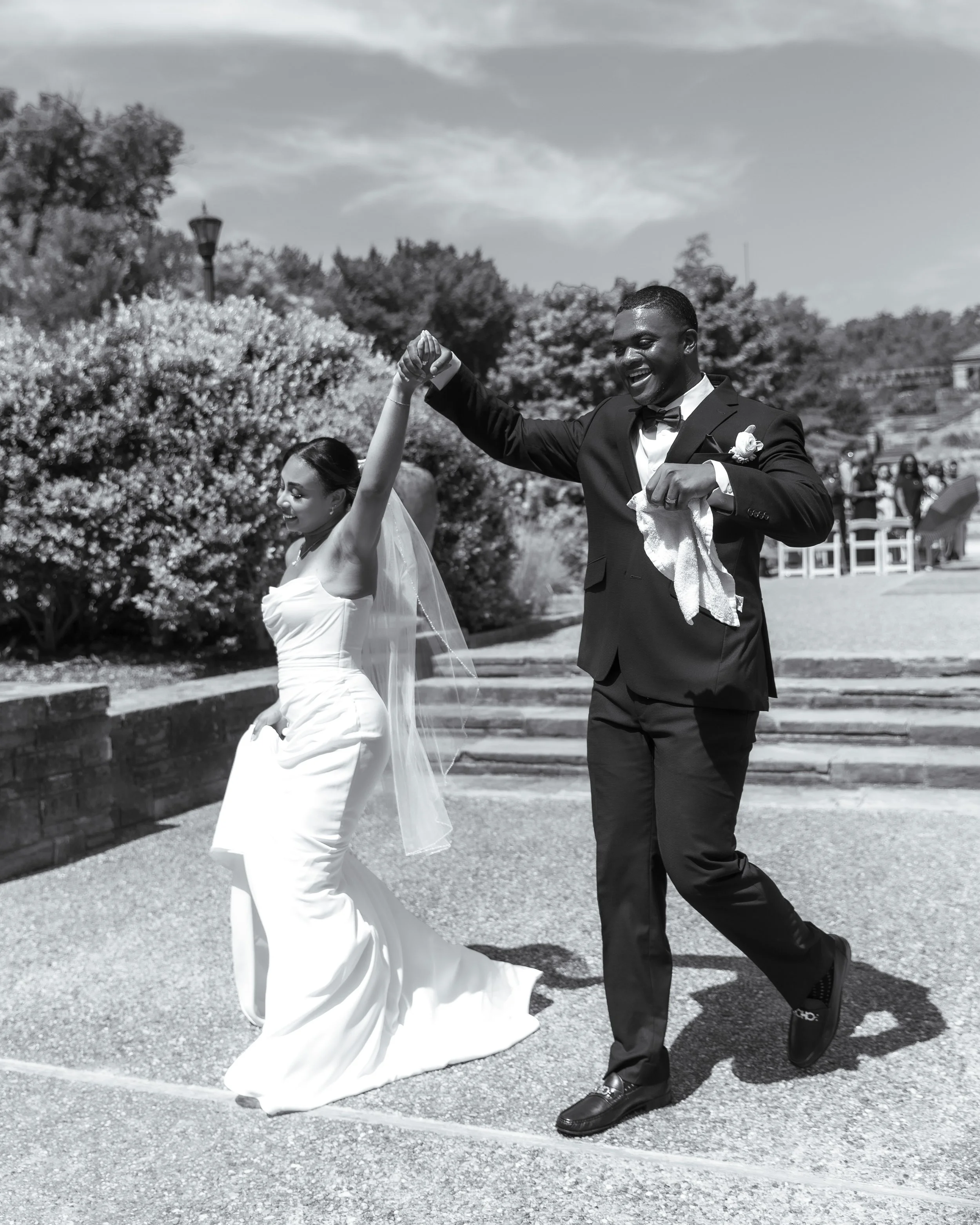 A bride and groom dancing outside on their wedding day, with the groom lifting the bride's hand, in black and white.