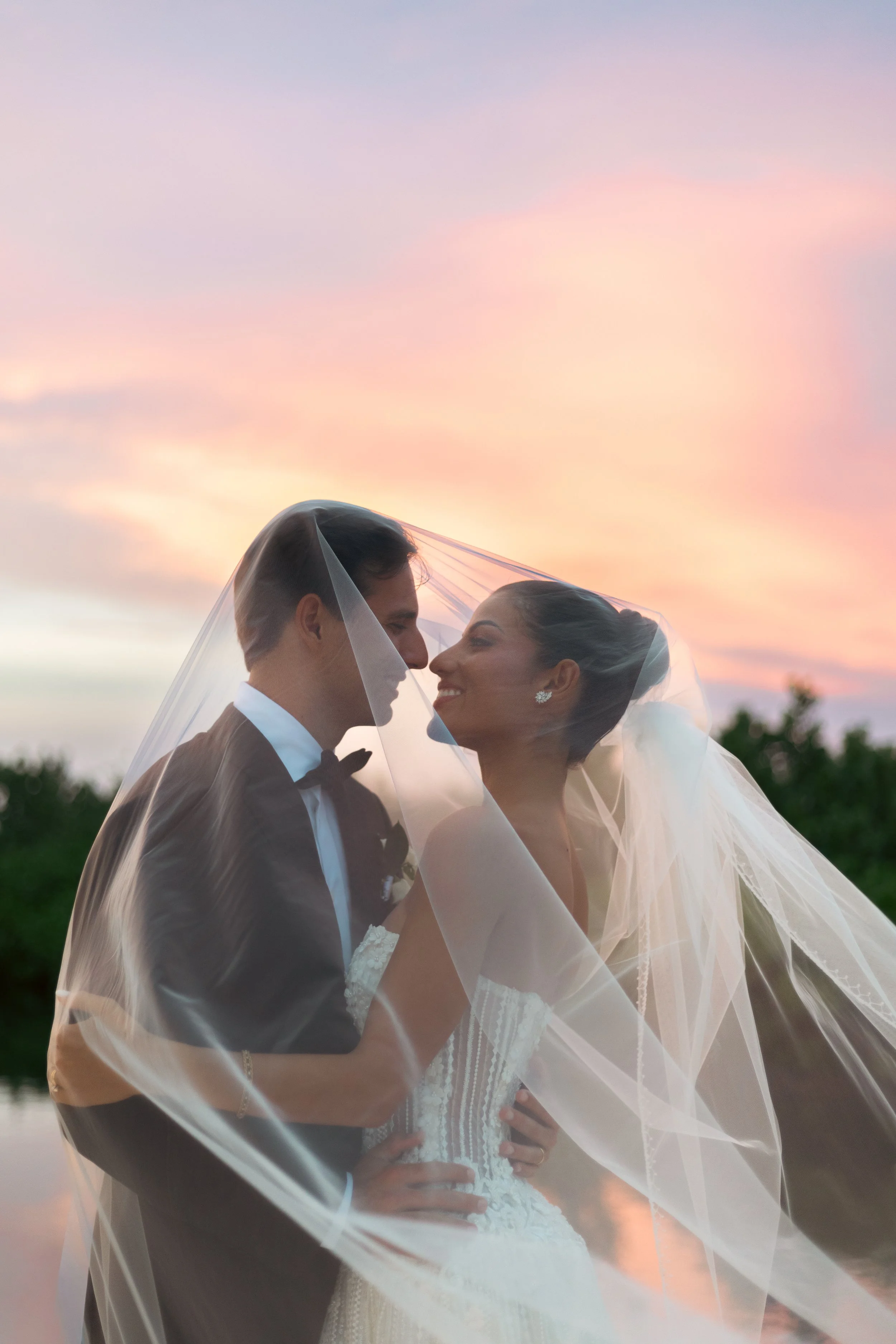 A bride and groom smiling and close together, standing outdoors at sunset under a veil.