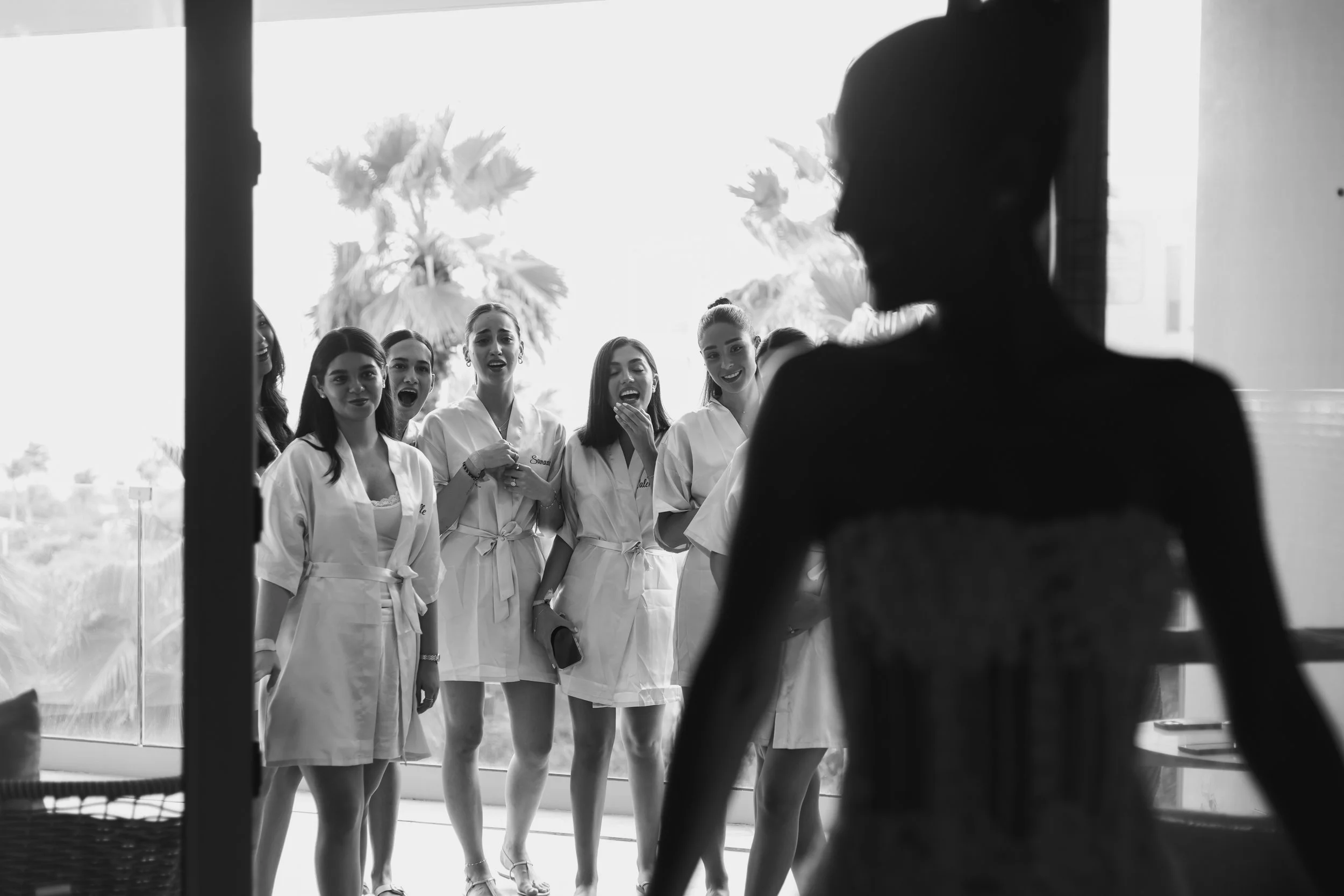 A group of women wearing robes, expressing surprise and happiness, with a woman in the foreground in silhouette, possibly getting ready for a wedding or special event.