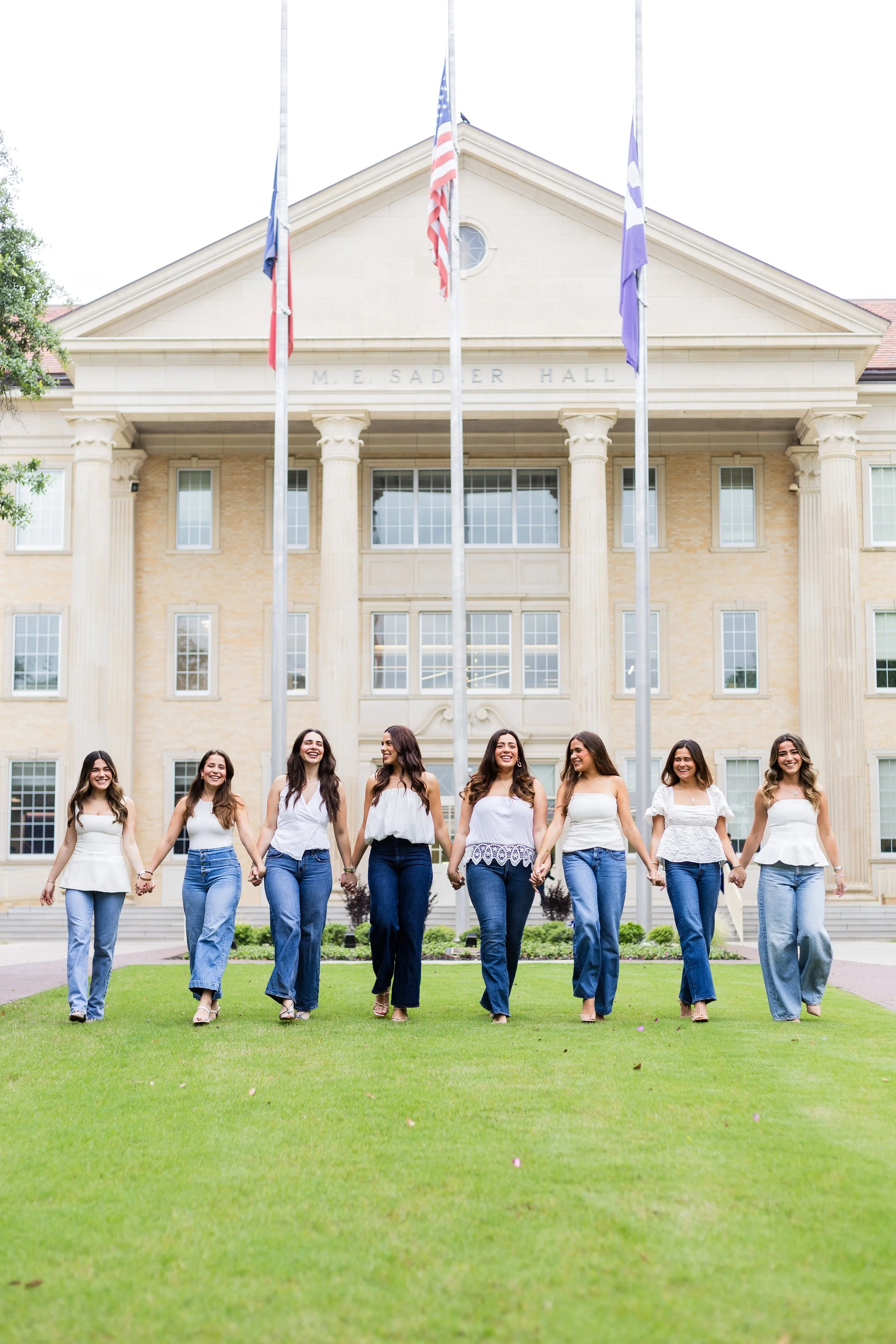 Group of seven women holding hands and walking on a well-maintained lawn in front of a classical-style building with columns and flags.