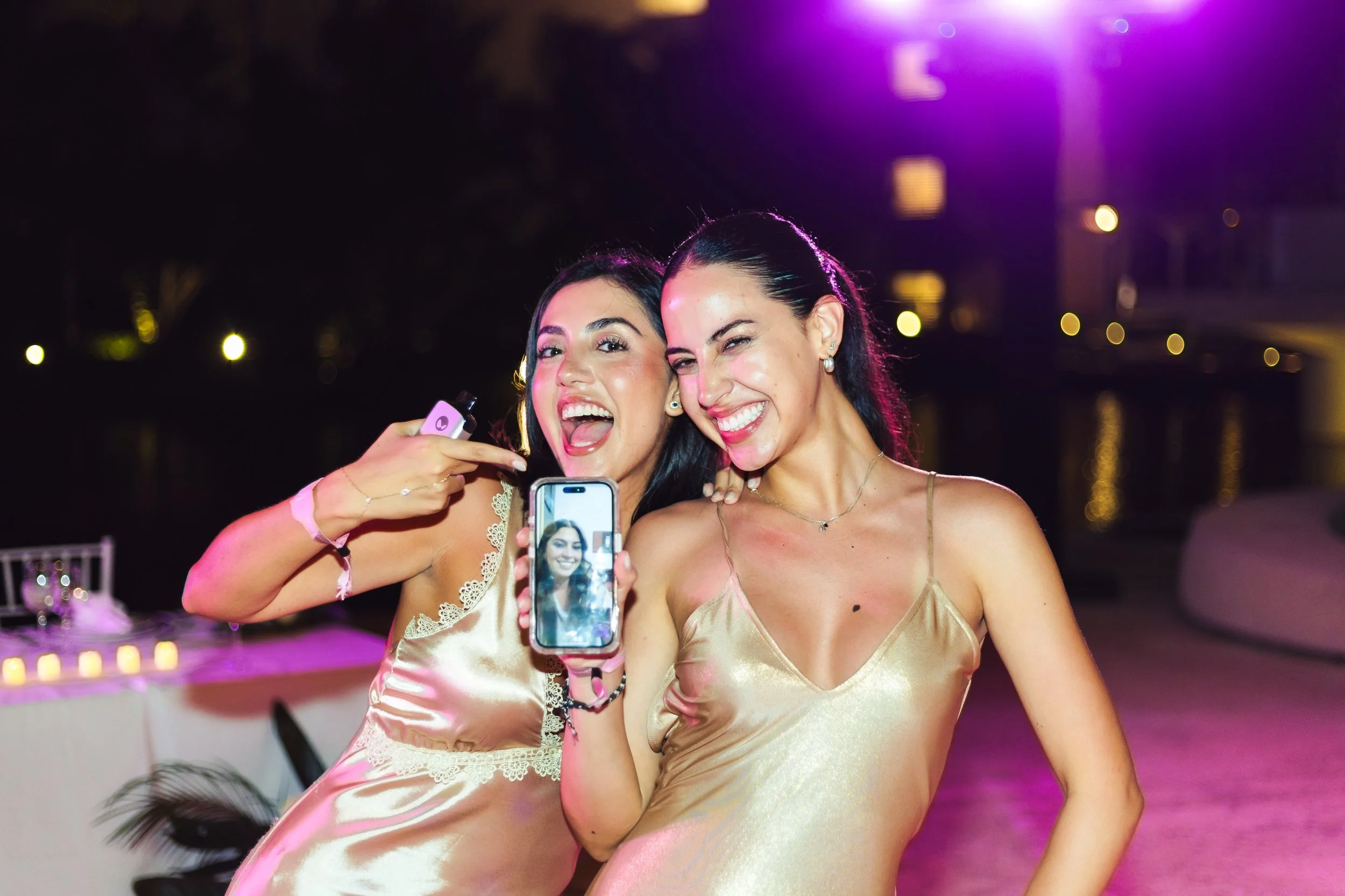 Two women in gold satin dresses taking a selfie at an outdoor nighttime event with purple lighting and a waterfront in the background.