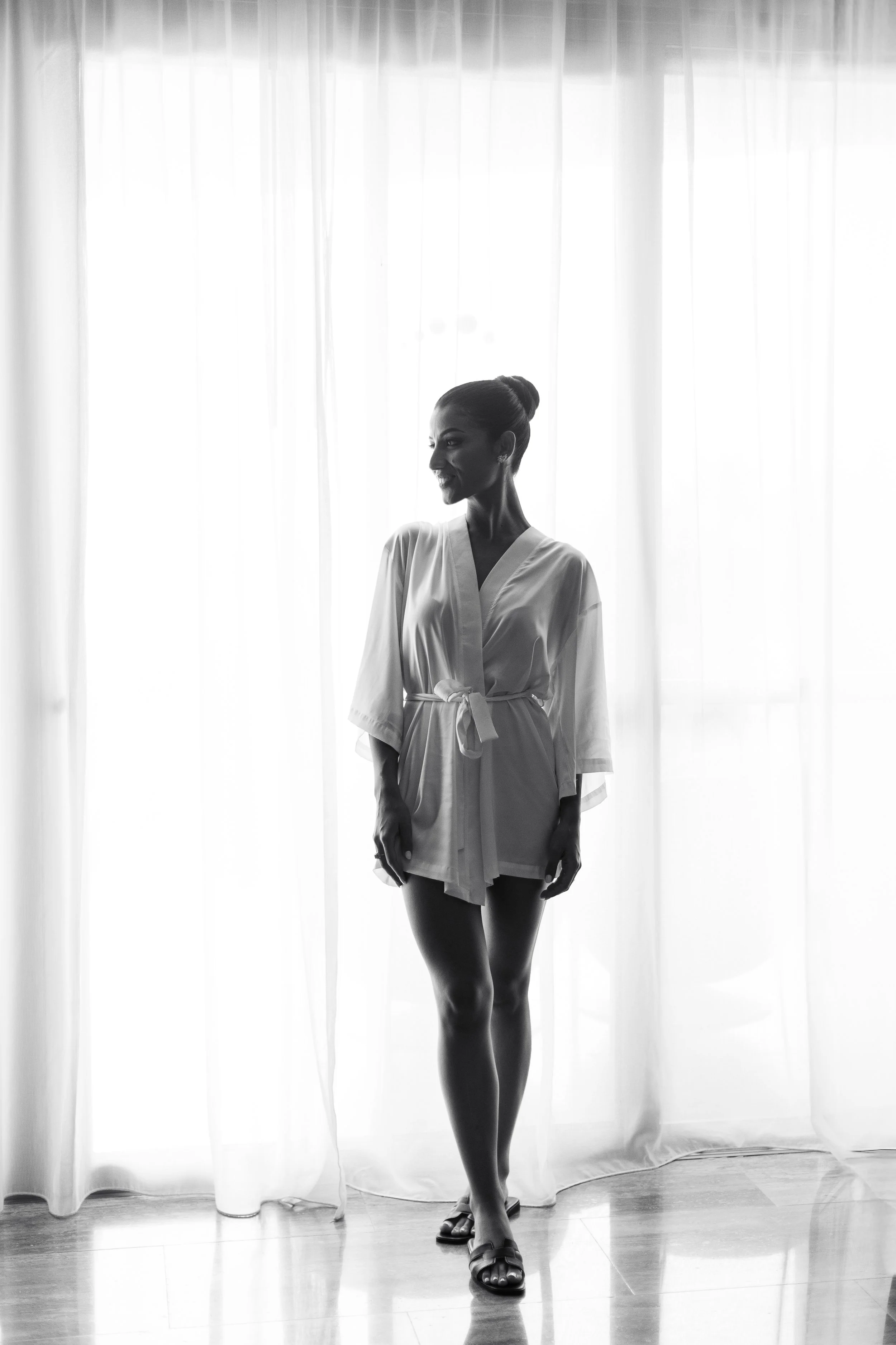 A woman standing by sheer curtains, wearing a robe and sandals, backlit in black and white.