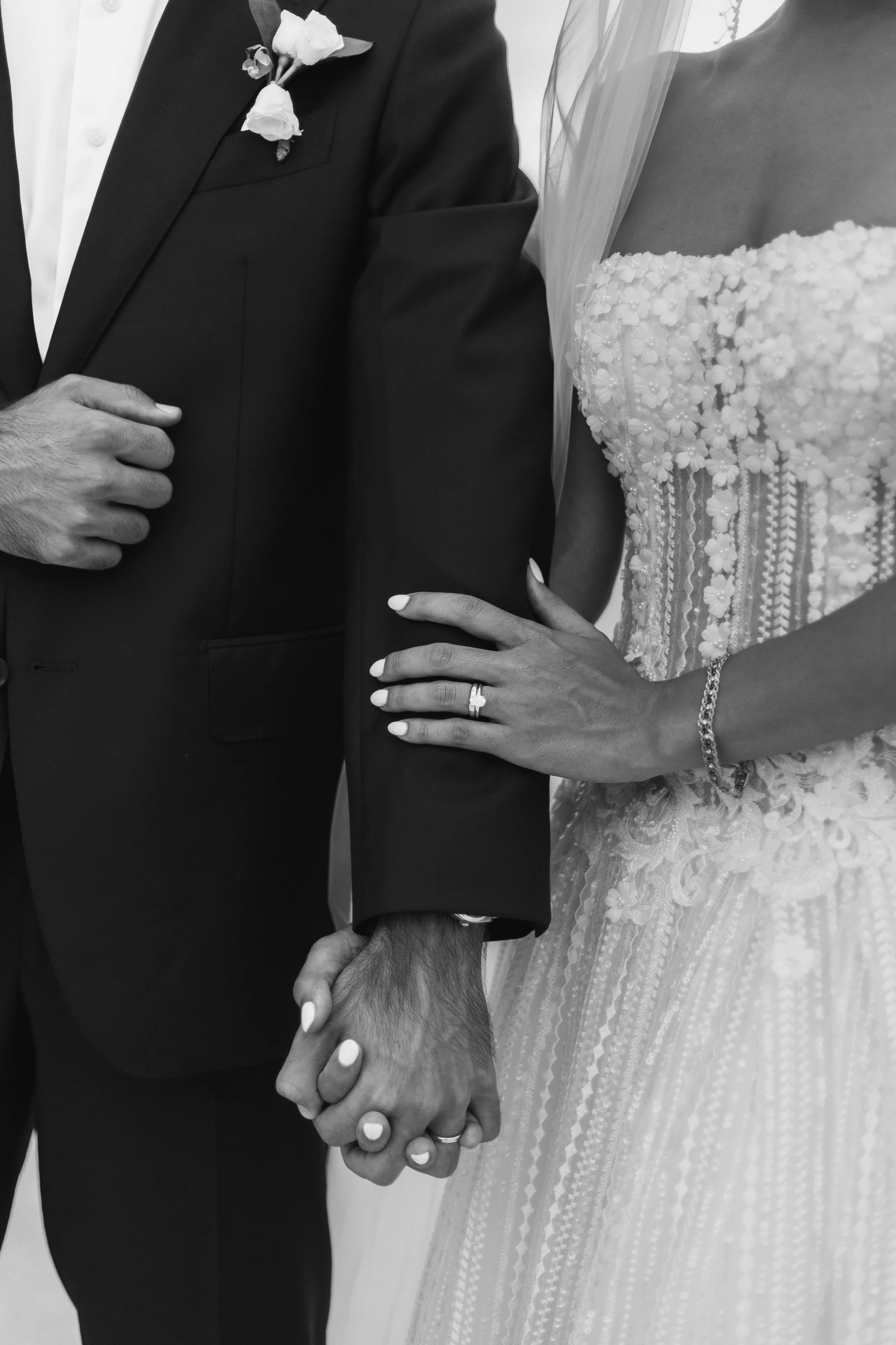 A black-and-white photo of a bride and groom holding hands, showing their wedding rings and attire.