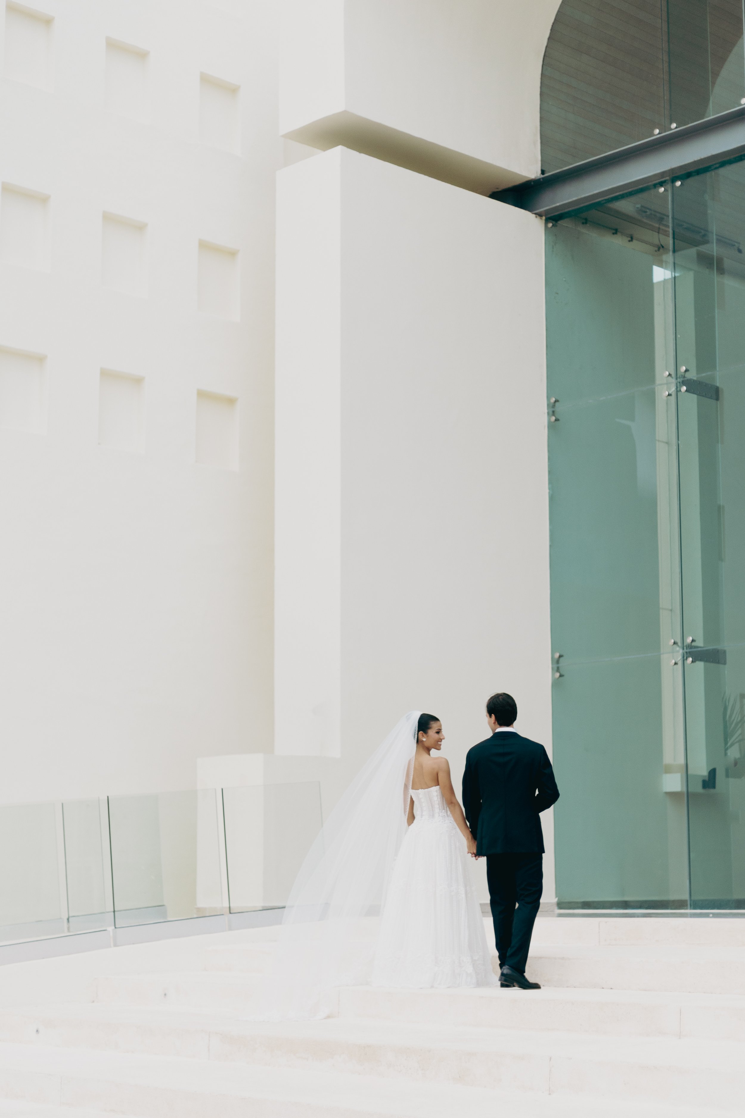 A bride and groom holding hands and walking outdoors near a modern white building with glass windows.