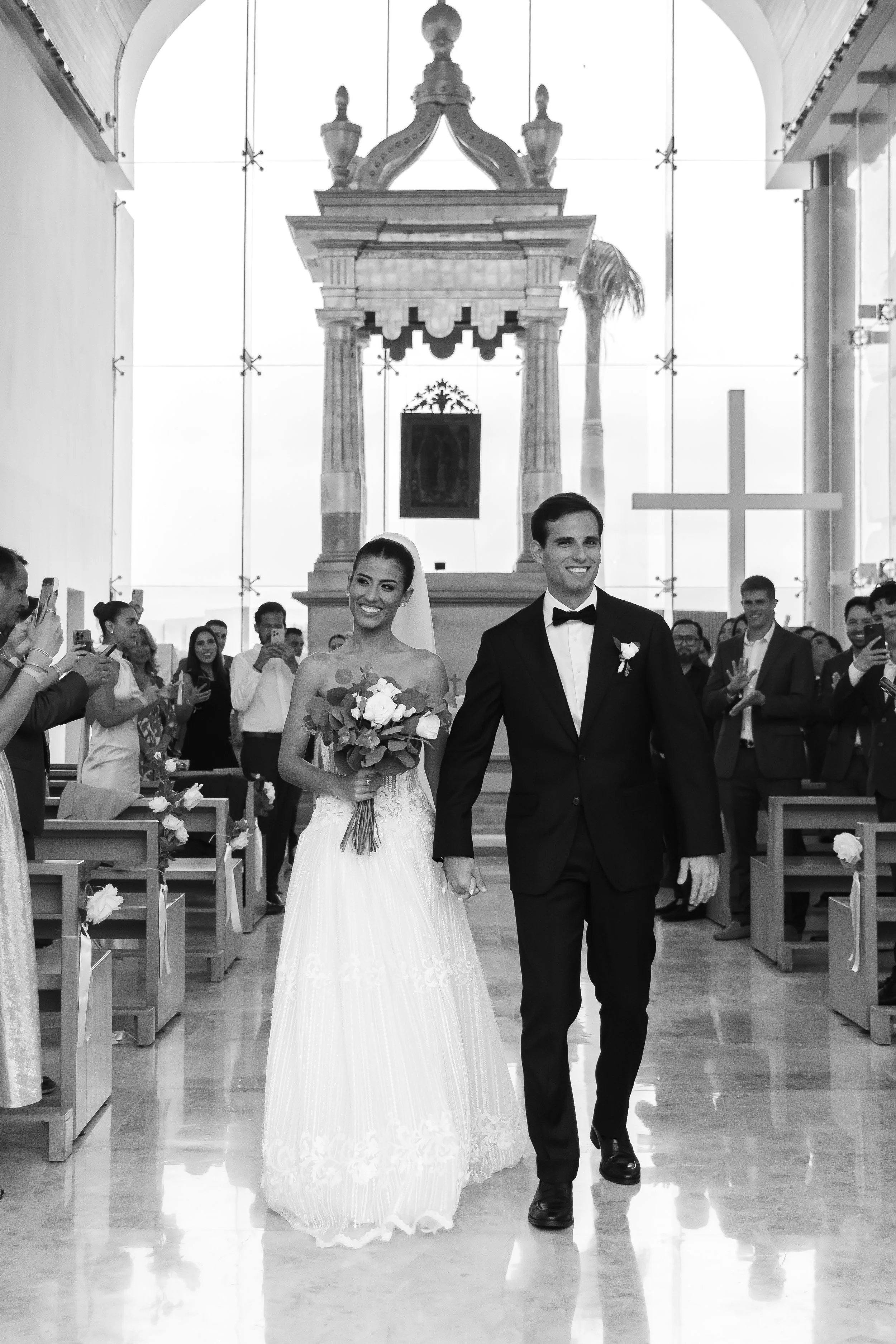 Black and white photo of a bride and groom walking down the aisle inside a church, surrounded by guests taking pictures.