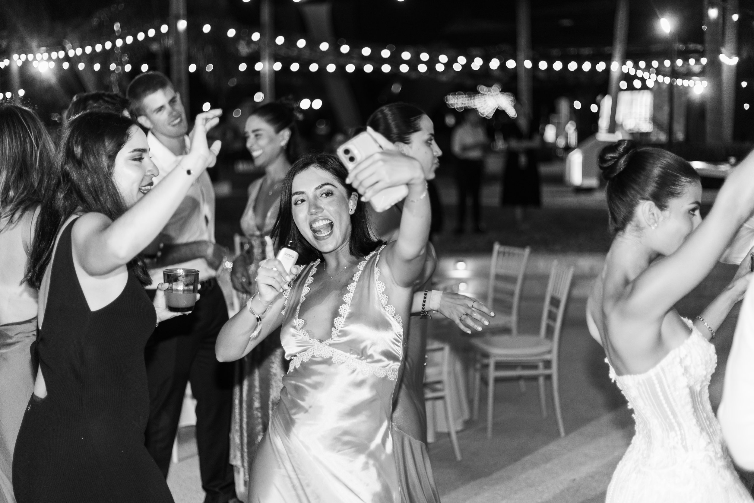 A group of people dancing and taking photos at a nighttime party with string lights overhead.