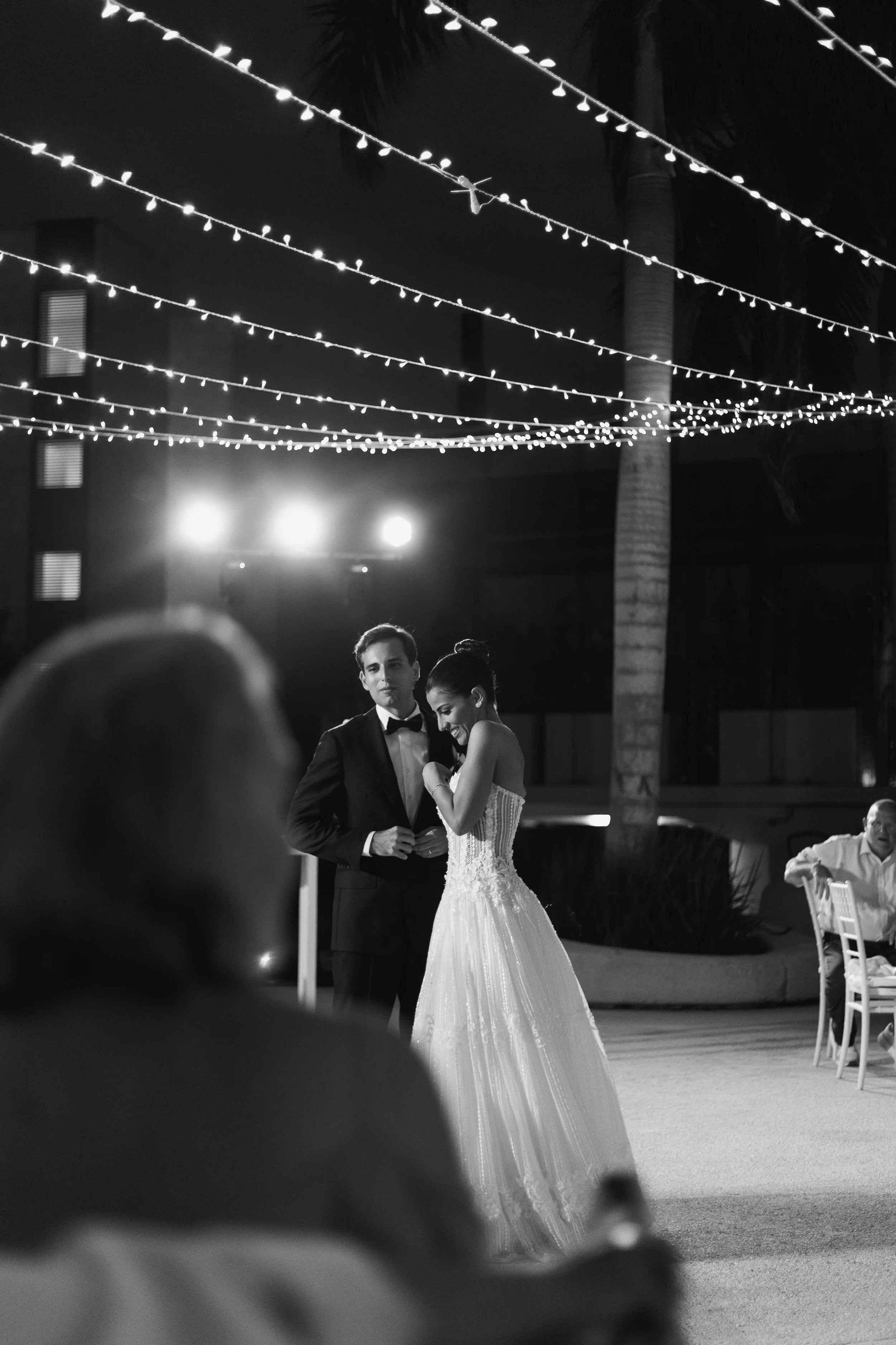 A black and white photo of a couple in formal attire dancing at a wedding reception outdoors at night. String lights are hanging overhead, and there are palm trees in the background. One guest is visible in the foreground and another guest sitting at