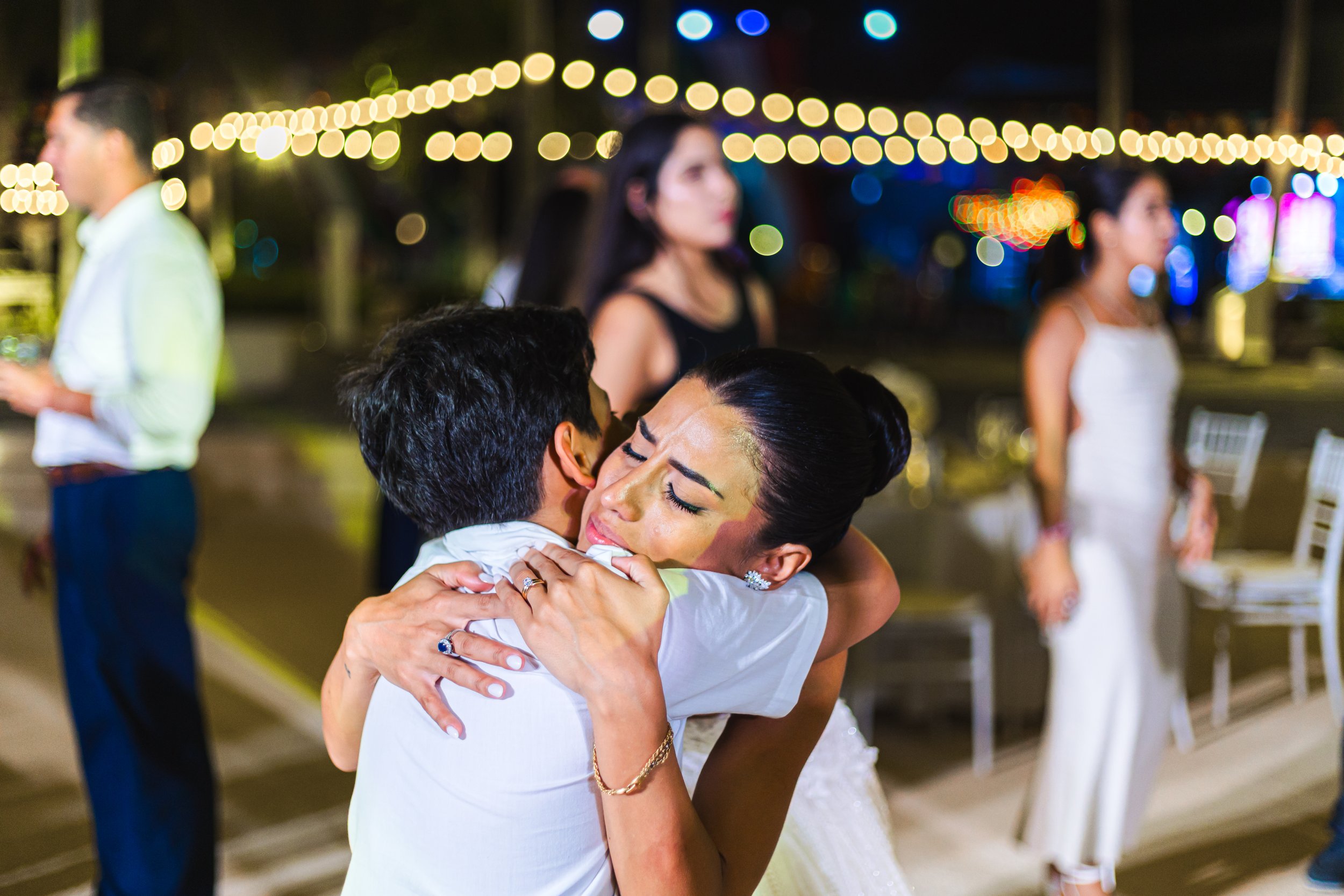 A woman in a white dress and a man in a white shirt are hugging at an outdoor event at night with string lights overhead. Several other people can be seen in the background, including a woman in a black dress and a woman in a white dress, all under f