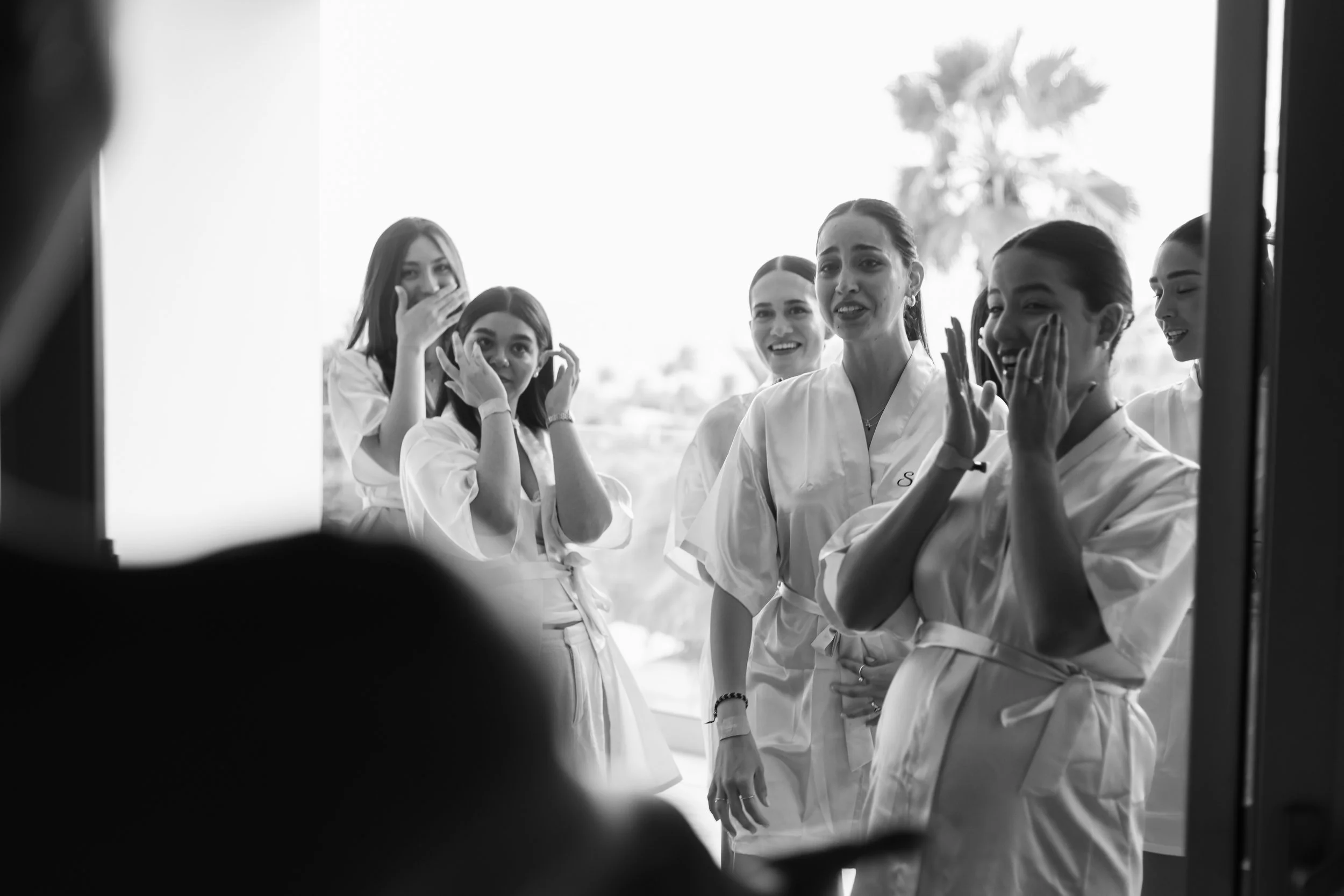 A group of six women in bridal robes appears emotional and surprised as they see someone, possibly the bride, on the other side of a glass door, with palm trees visible in the background.