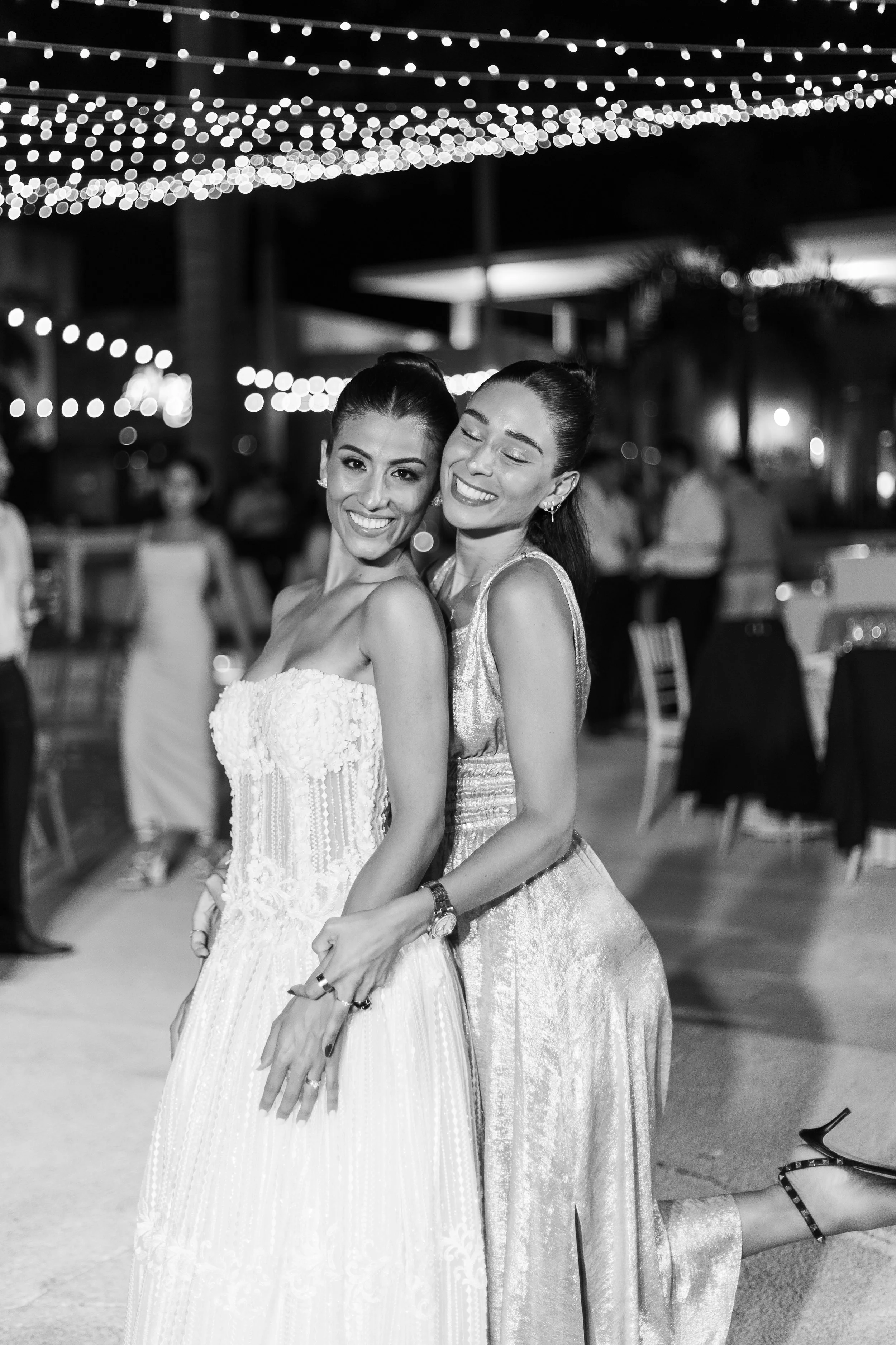 Two women in formal dresses smiling and hugging at an outdoor event with string lights overhead