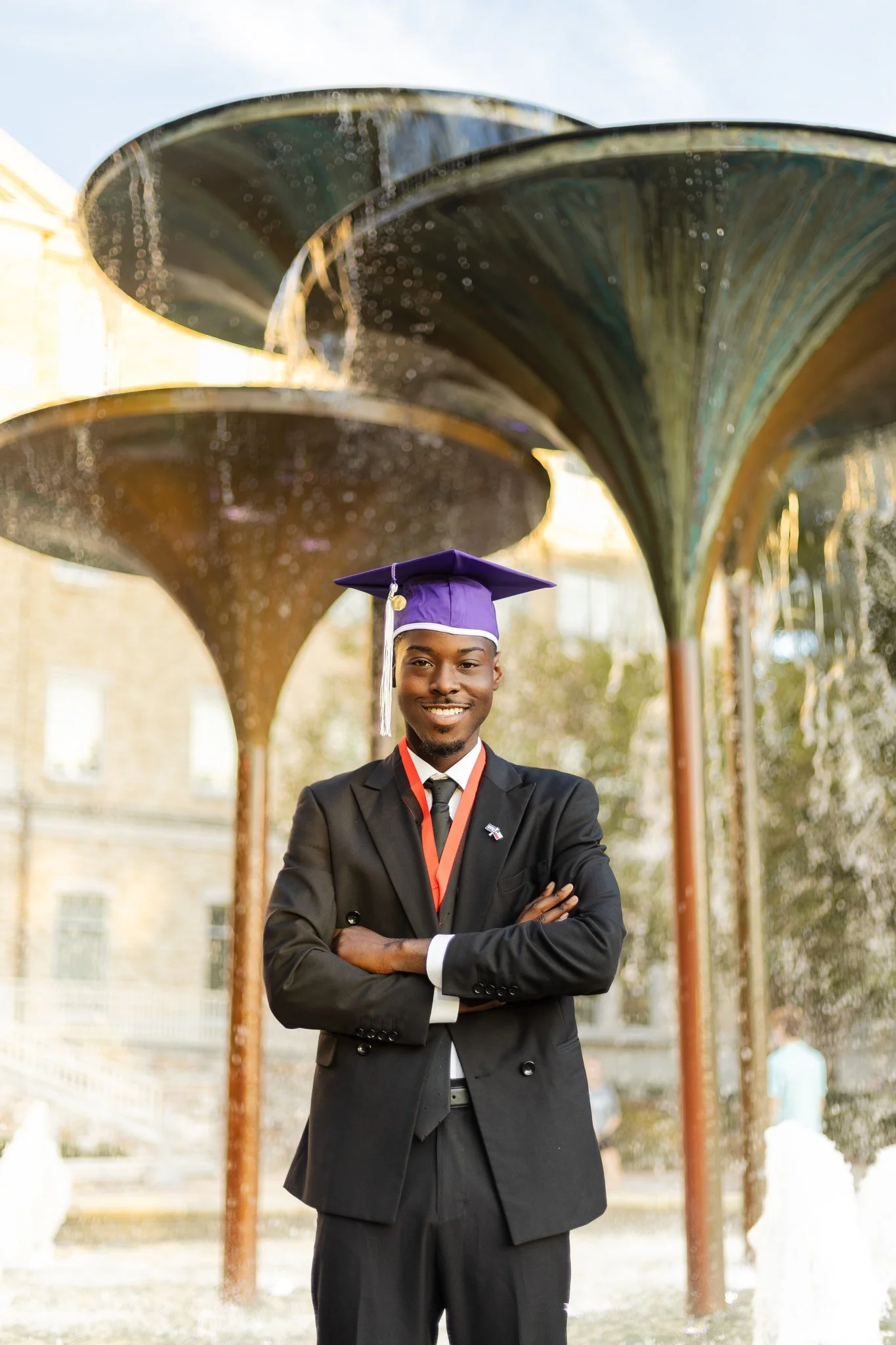 Young man in a suit and purple graduation cap standing in front of a fountain, smiling with arms crossed.