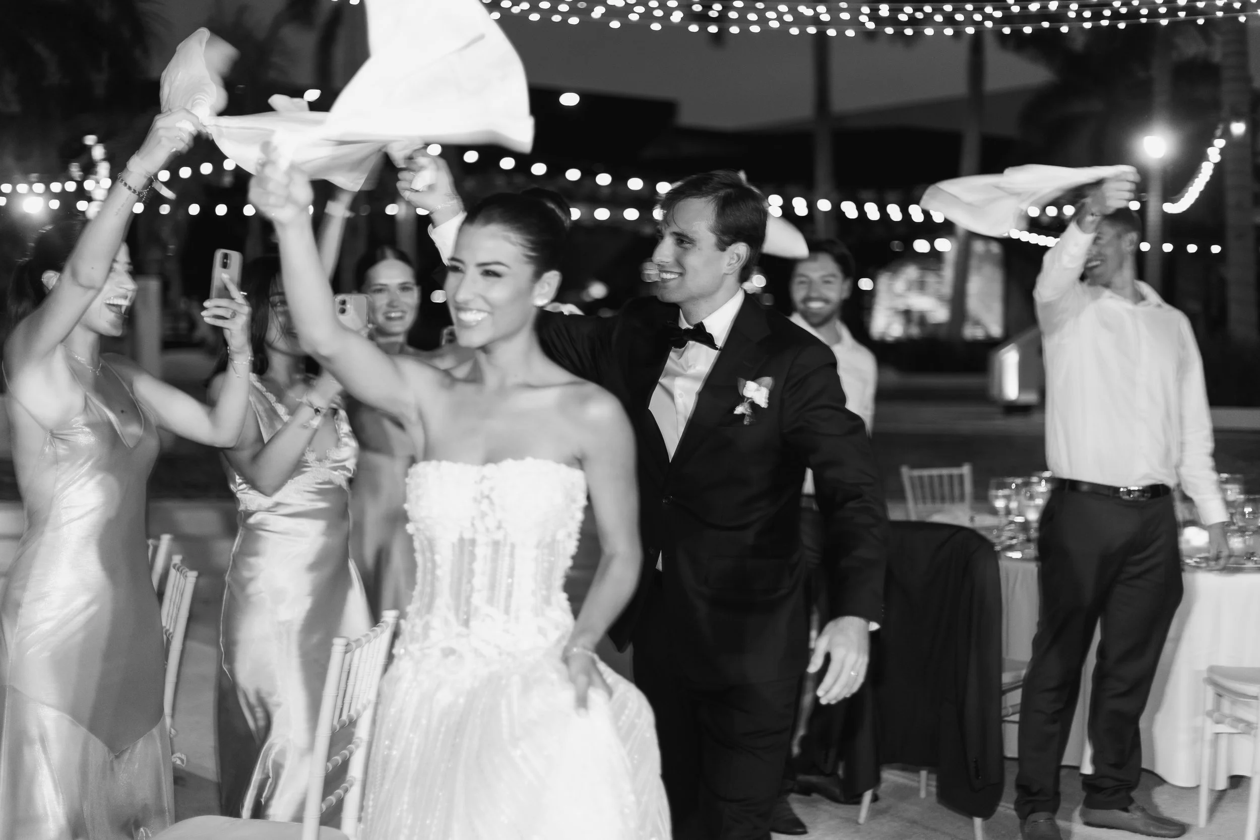 A bride and groom celebrating with wedding guests at a reception, dancing and waving napkins under string lights in an outdoor setting.
