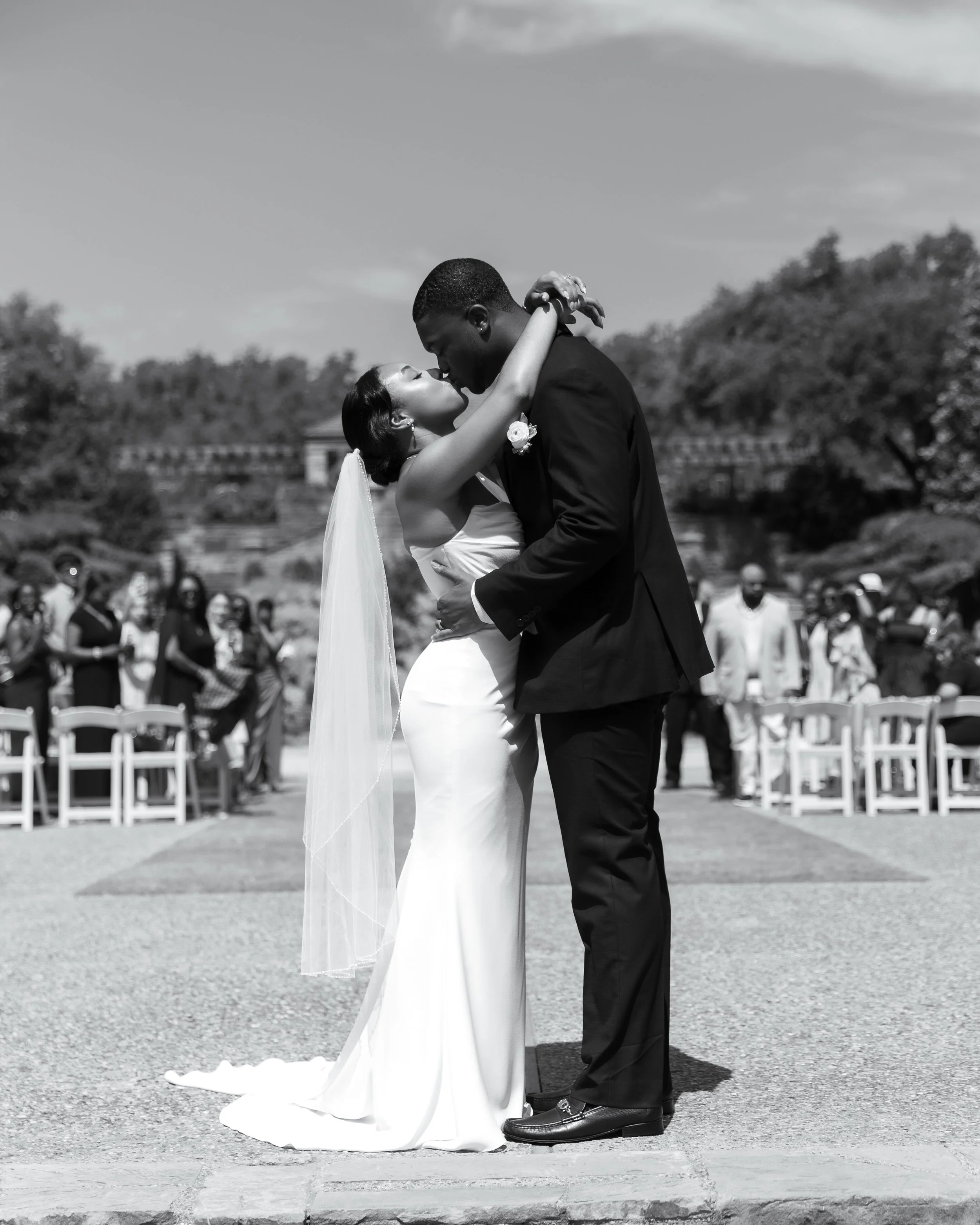 A black-and-white photo of a bride and groom kissing during their wedding ceremony outdoors, with guests and trees in the background.